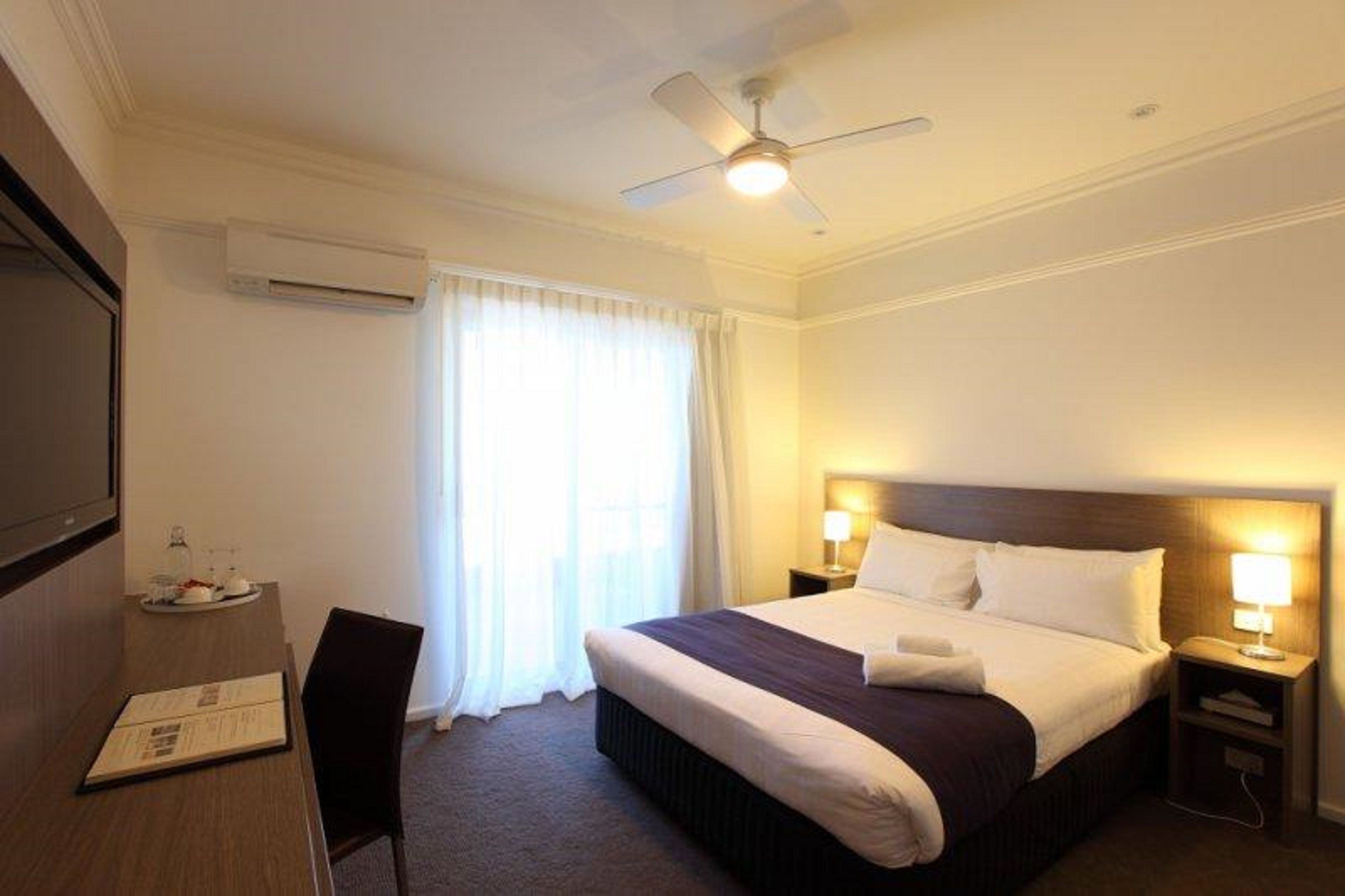 Een hotelkamer met een bed, een bureau met stoel, een plafondventilator en een raam met gordijnen.