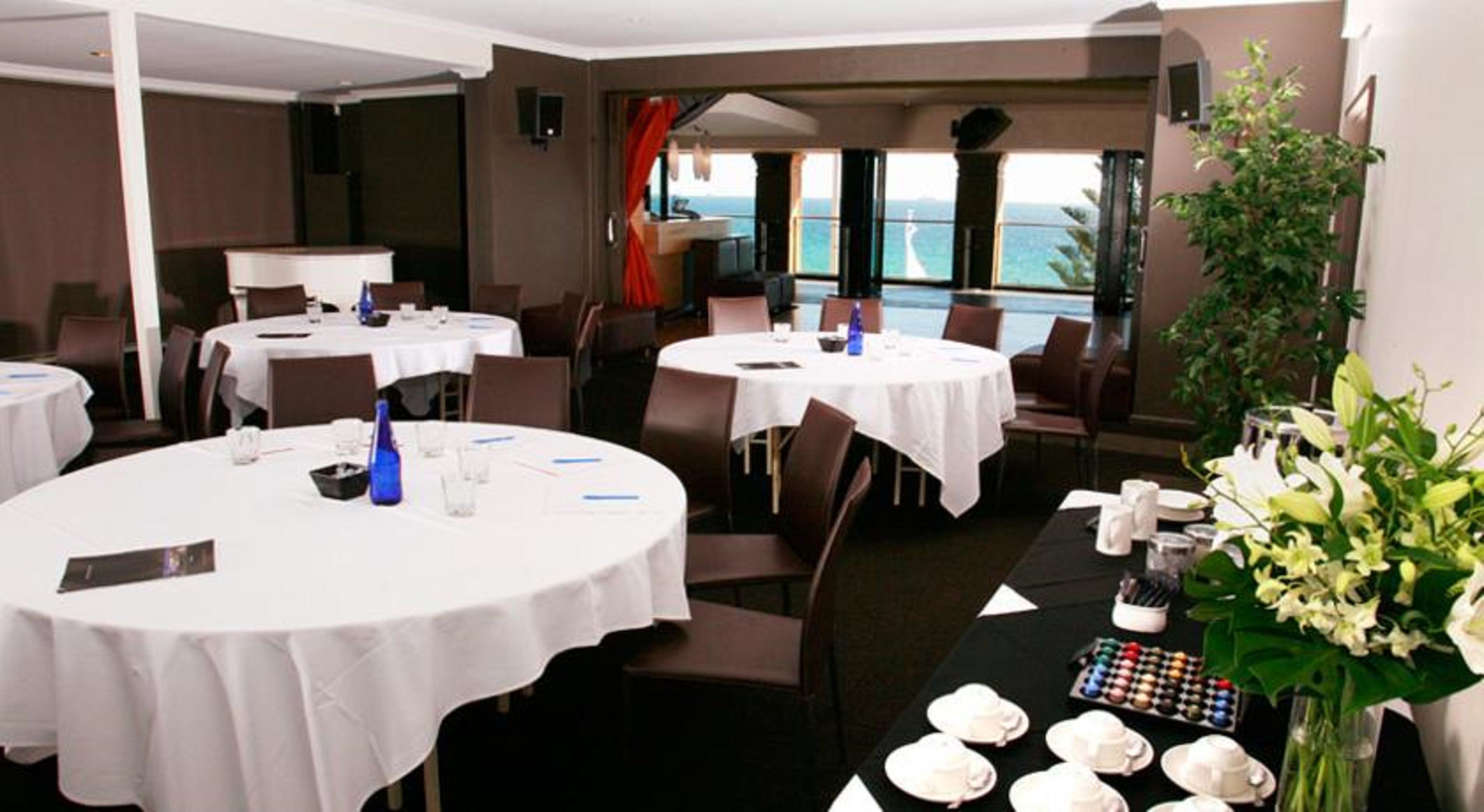 Un comedor con mesas redondas dispuestas para una comida, una mesa de buffet con diversos platos, y vistas al océano a través de amplias ventanas.