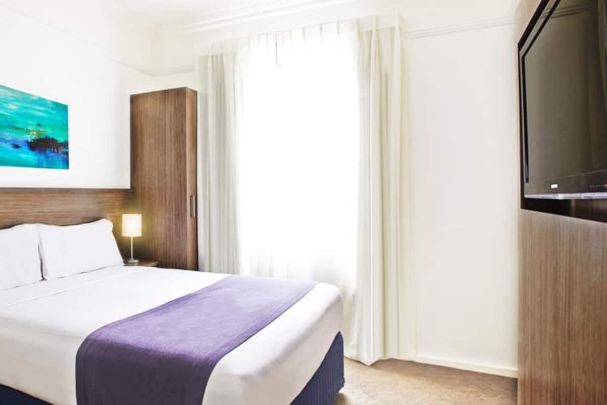 Et hotelværelse med en seng, et natbord, en lampe, et billede, et klædeskab og et fjernsyn.