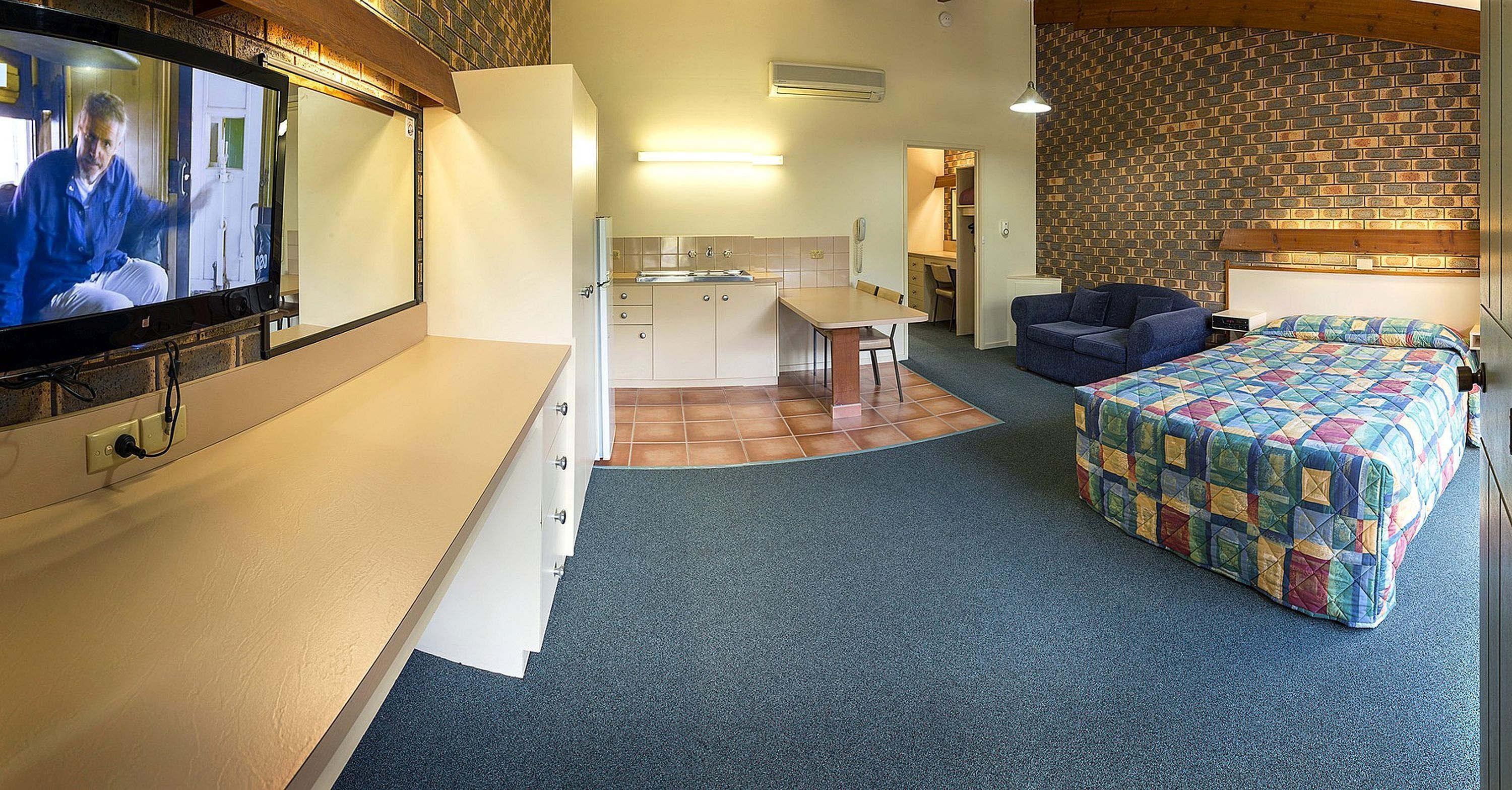 A compact hotel room with a kitchenette, a bed with a colorful quilt, a seating area with a blue sofa, and a television mounted on the wall.