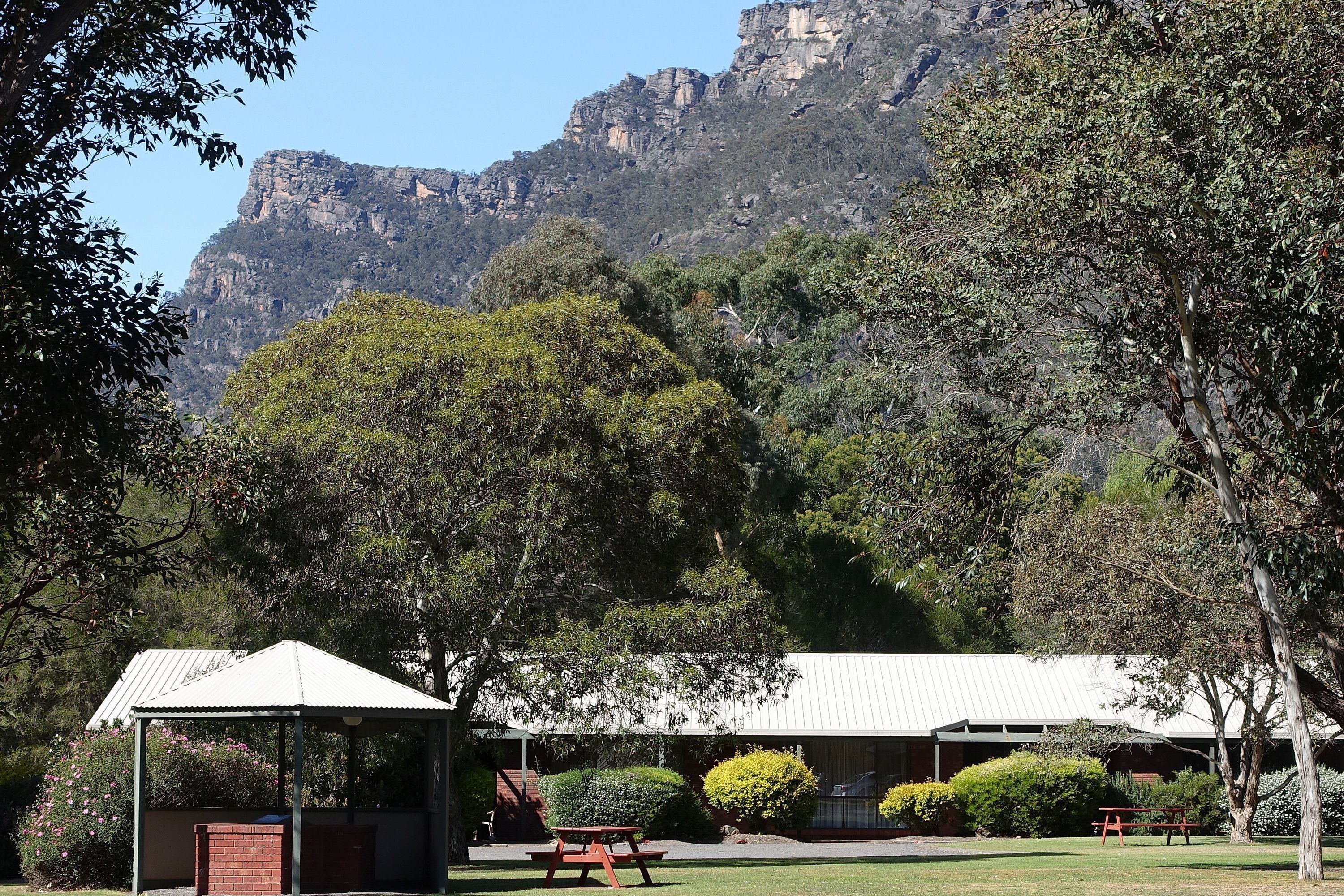 A pavilion with a roof, surrounded by trees and a mountain backdrop.
