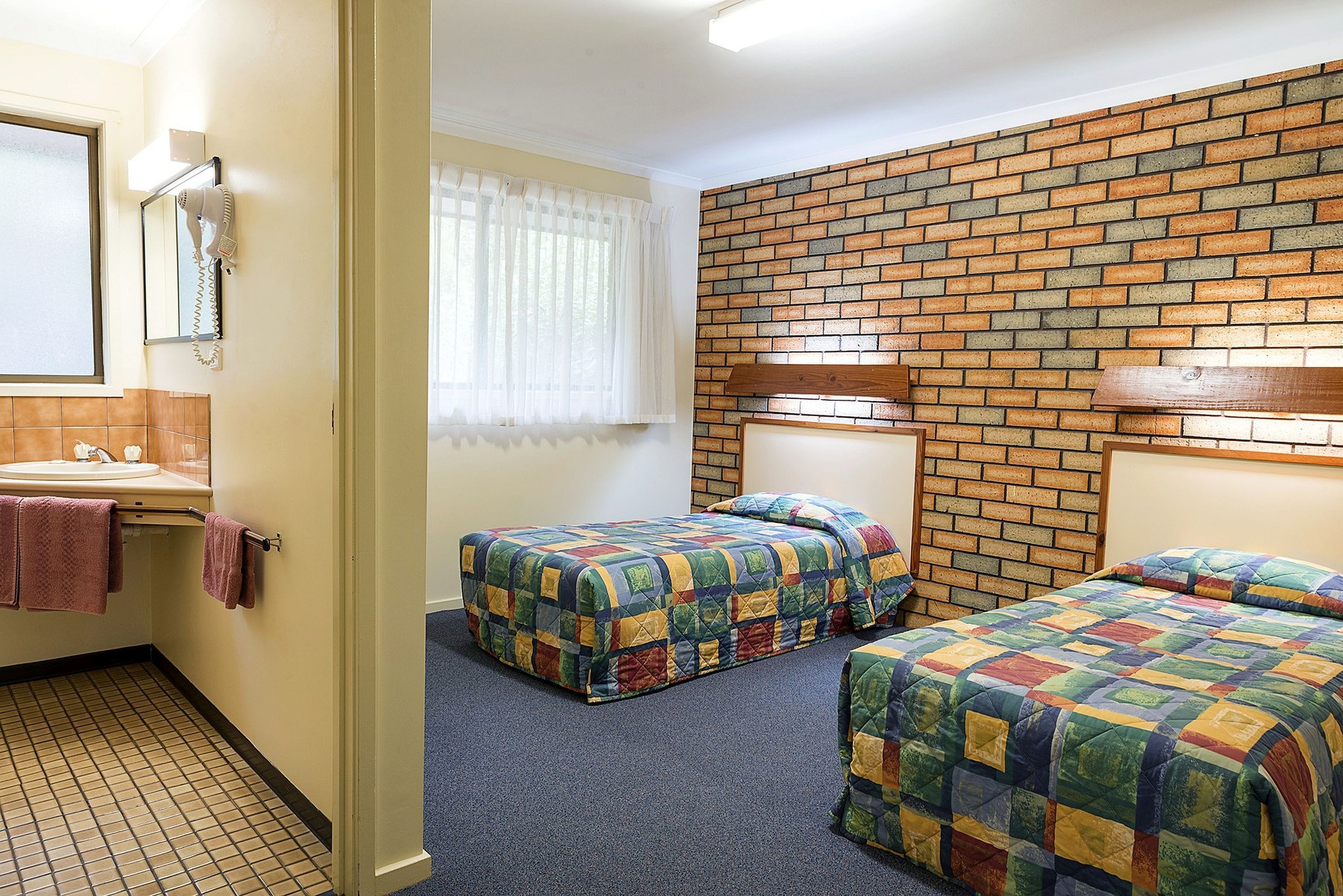 A hotel room with two beds, a bathroom with a sink, and a brick-patterned wall.