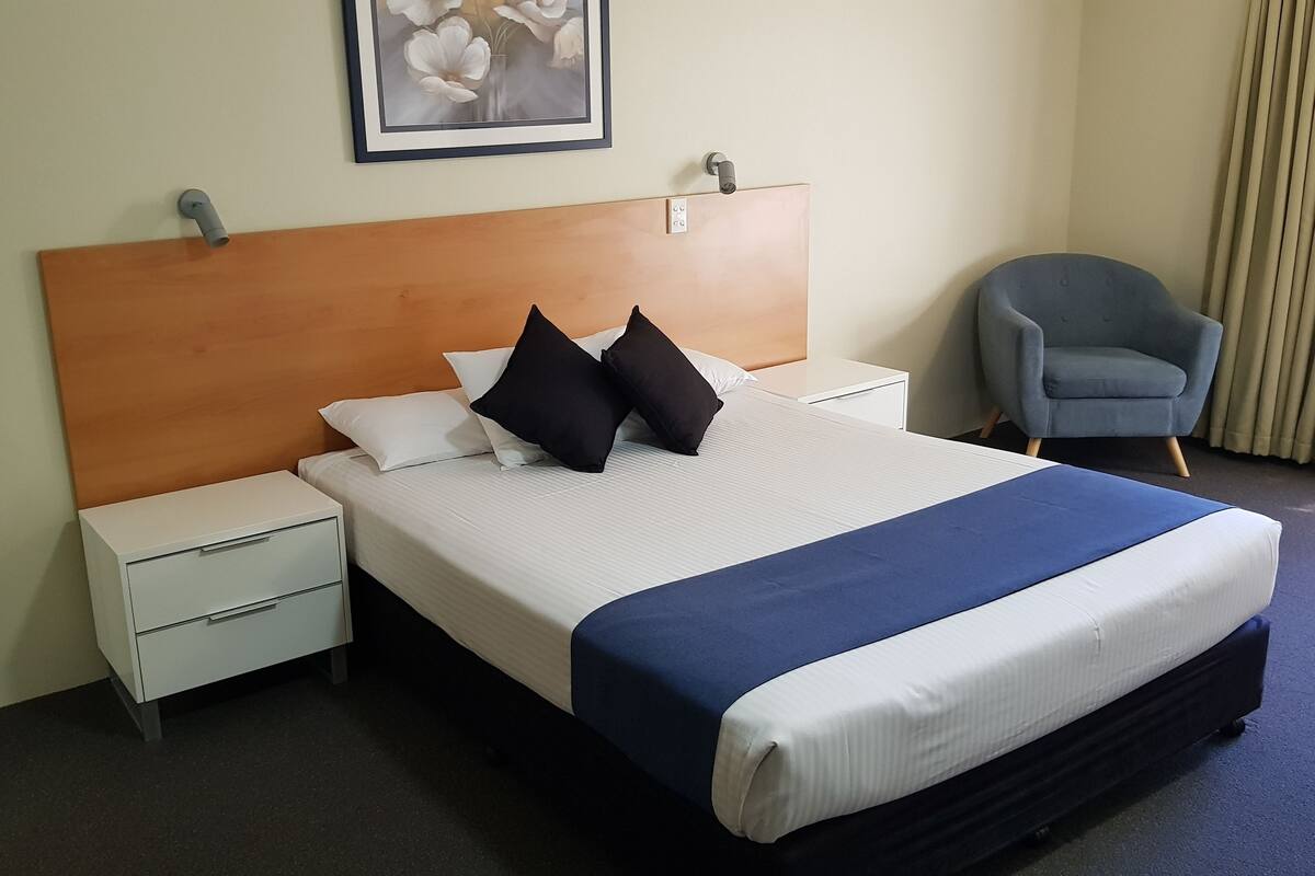 Hotellihuone, jossa on sänky, yöpöytä, tuoli ja seinälle ripustettu taulu.