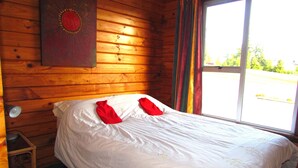 Una habitación de cabaña de madera con una cama, un taburete de madera y un ventanal con cortinas.