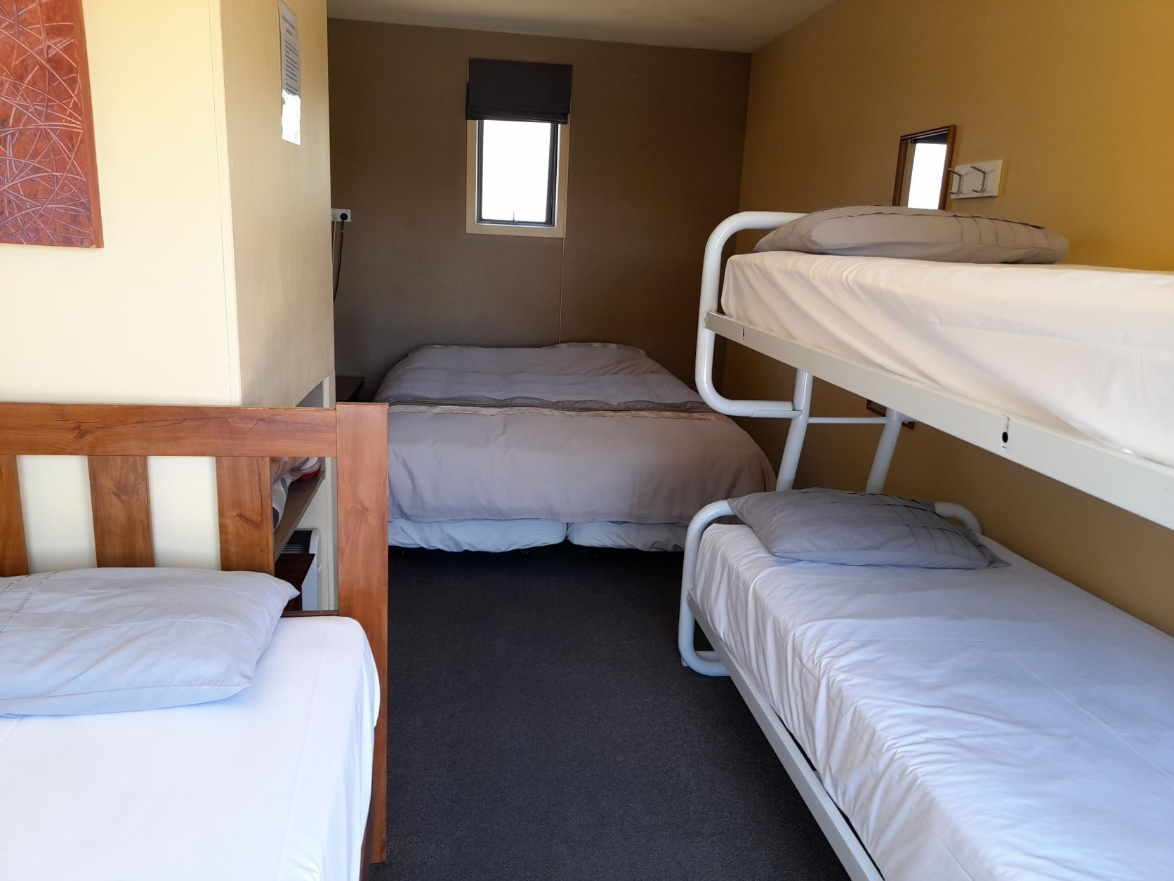 Habitación pequeña y funcional con literas y una cama individual. Tiene una ventana con cortina, un radiador y un espejo en la pared.