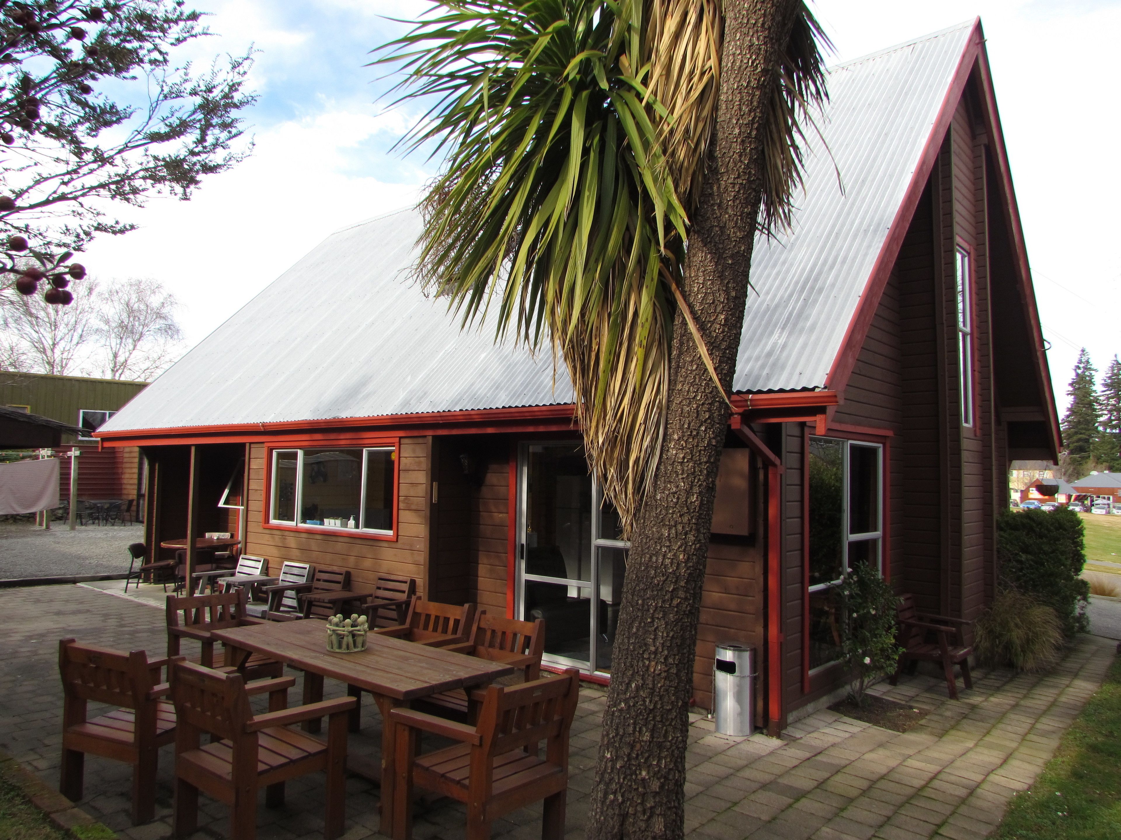 A wooden building with a sloped roof, a patio with wooden furniture, and a palm tree.