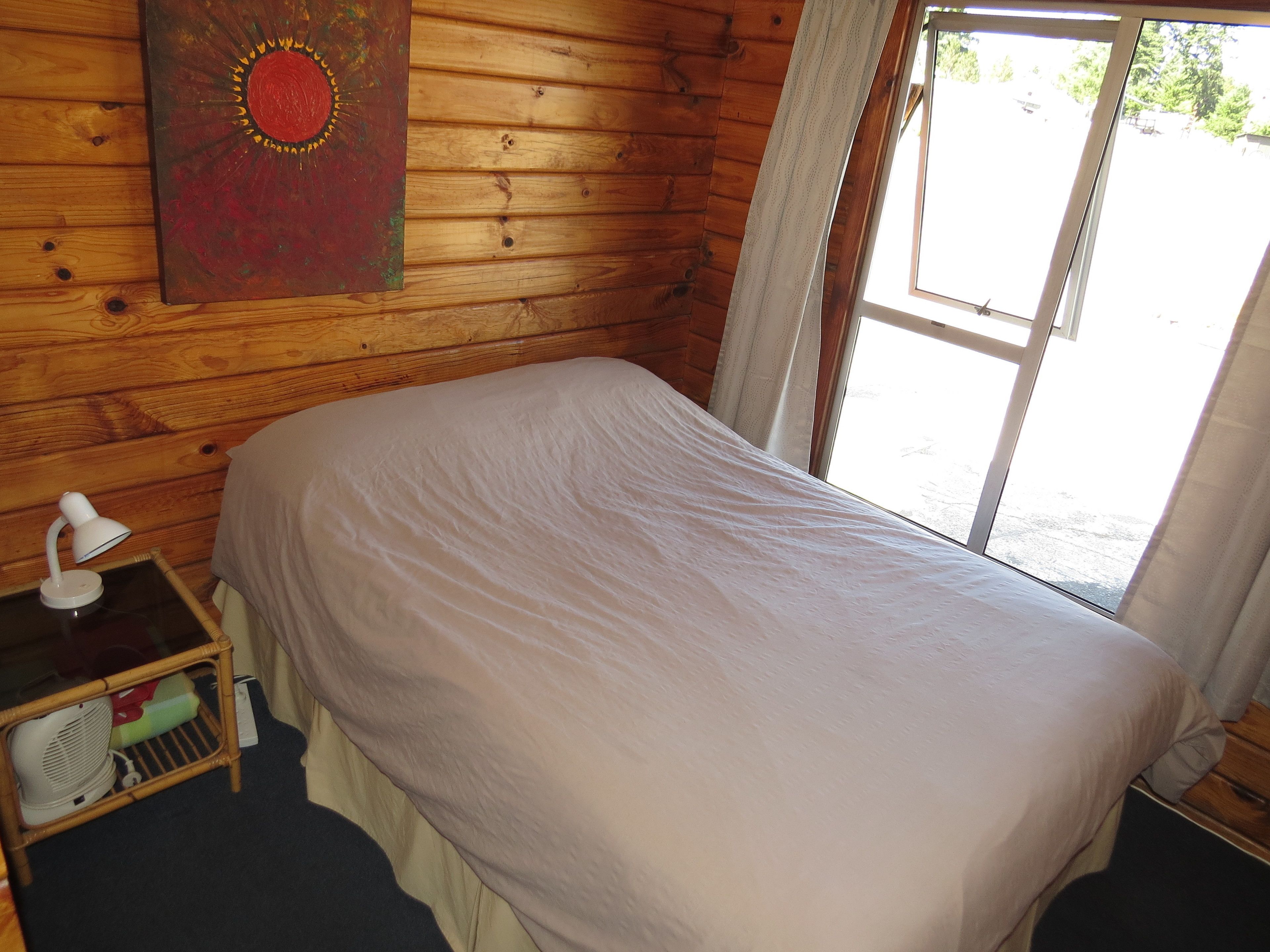 A wooden cabin room with a bed, a nightstand, a lamp, and a window with curtains.