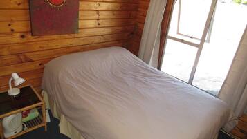 A wooden cabin room with a bed, a nightstand, a lamp, and a window with curtains.