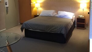 Hotellihuone, jossa on sänky, kaksi yöpöytälamppua, kattotuuletin, lasipöytä ja ovi, joka avataan avainkortilla.