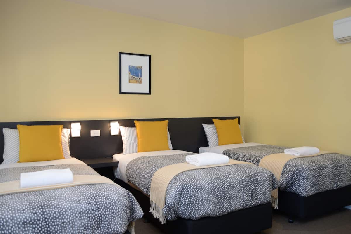 A hotel room with three beds, each with a patterned bedspread and a white towel. There is a framed picture on the wall above the beds.