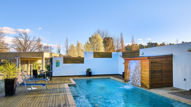 A swimming pool with a wooden deck, a small waterfall feature, and a covered seating area with chairs. - Motel On Alpine (Myrtleford)