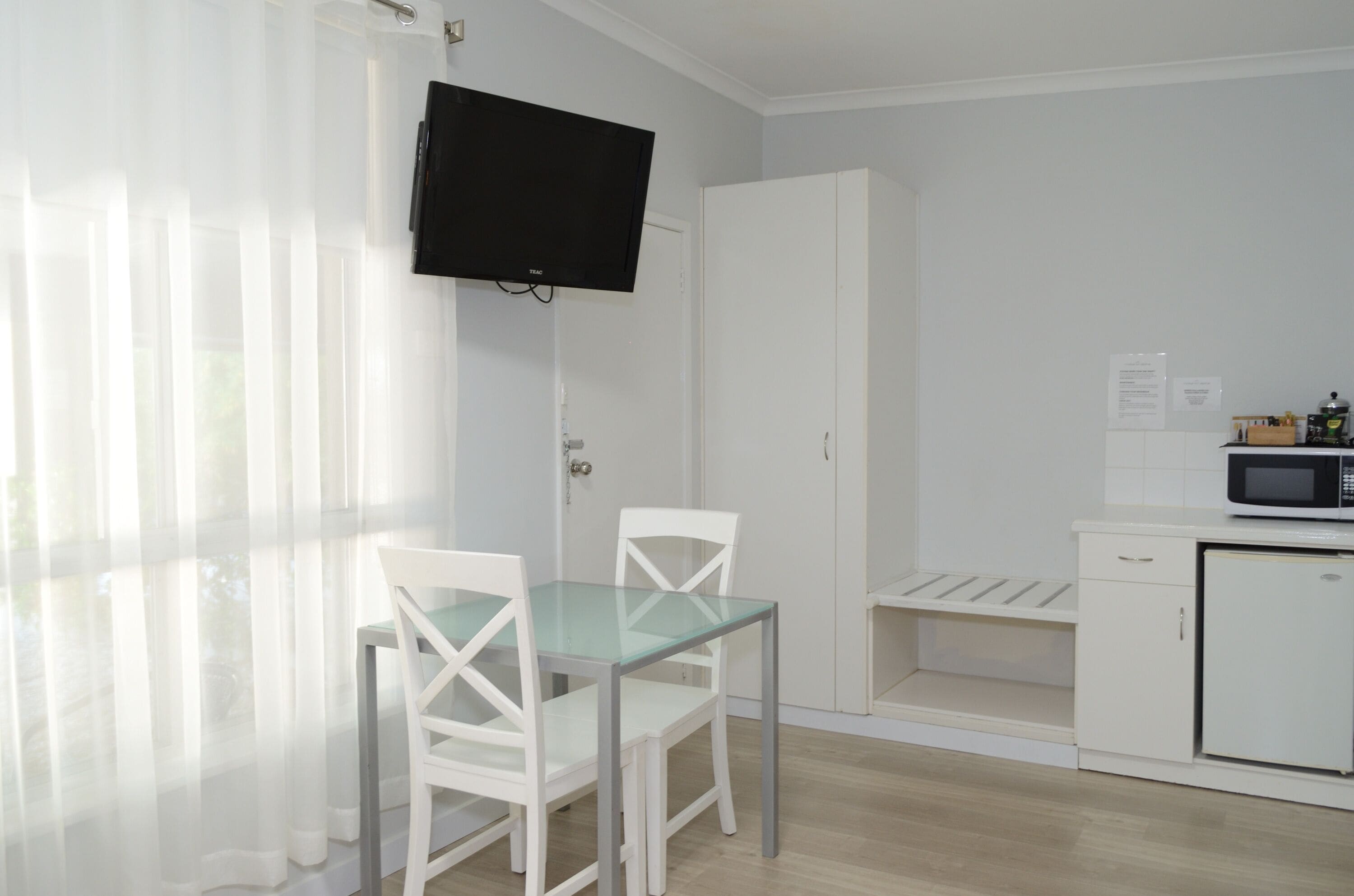 A small, bright room with a glass table and white chairs, a white wardrobe, a microwave, and a television mounted on the wall.