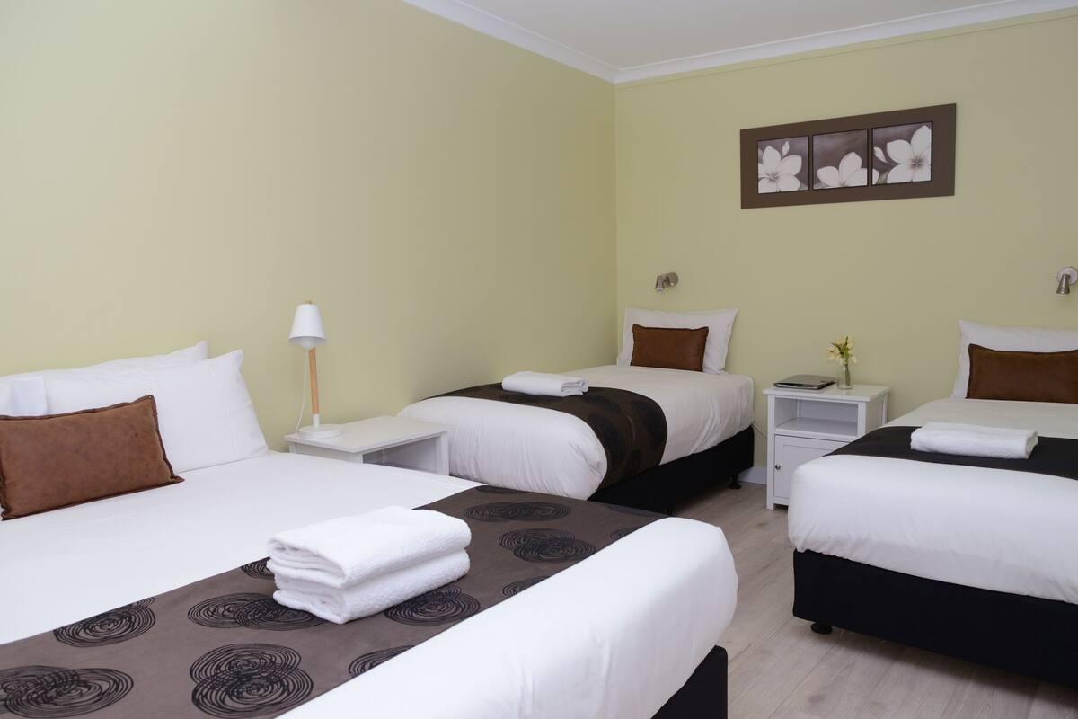 A hotel room with two beds, each with white linens and brown pillows. There are bedside tables with lamps and a small nightstand with a lamp. The walls are painted a light green, and there is a framed picture above the beds.