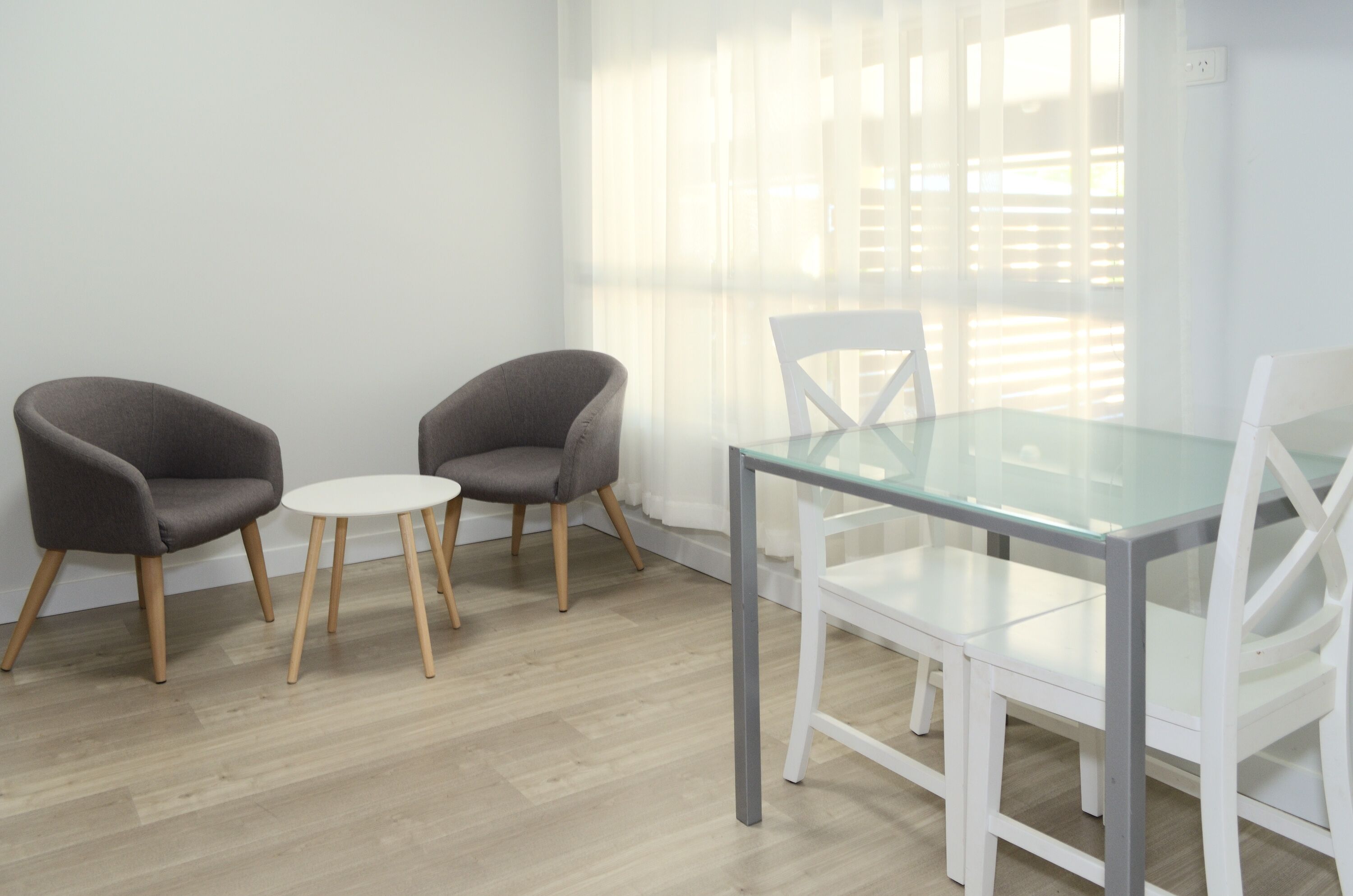 A room with a glass table and chairs, wooden flooring, and a large window with blinds.