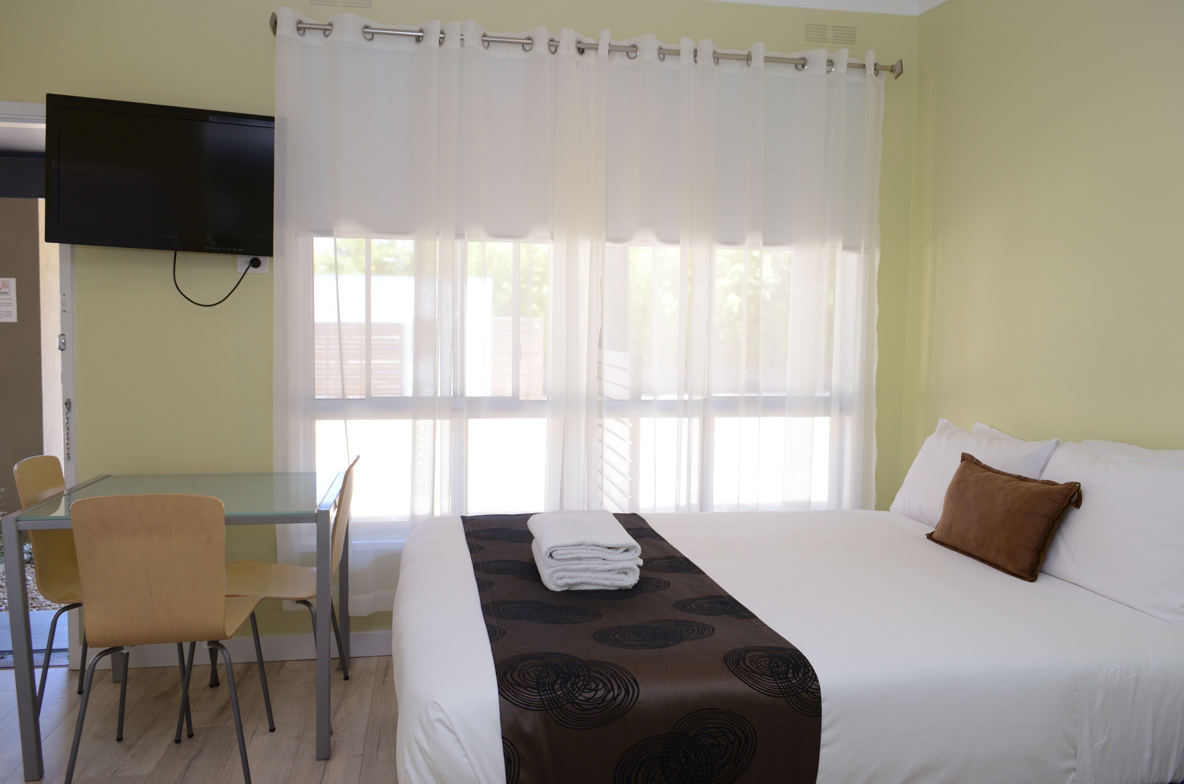 Ein Hotelzimmer mit Bett, Wandfernseher, Esstisch mit Stühlen und einem Vorhangfenster.