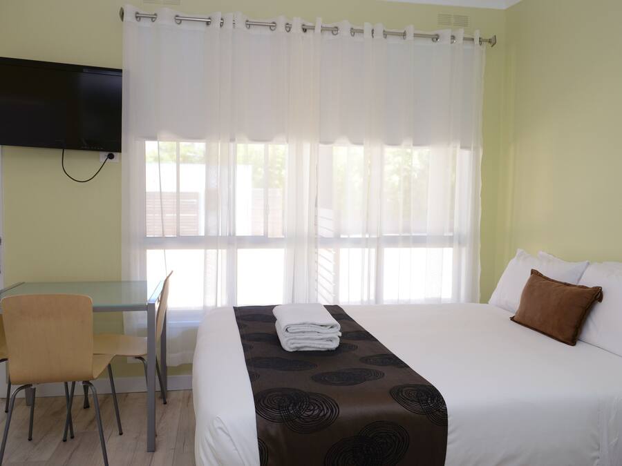 Habitación de hotel con cama, televisor en la pared, una mesa de comedor con sillas y una ventana con cortinas.