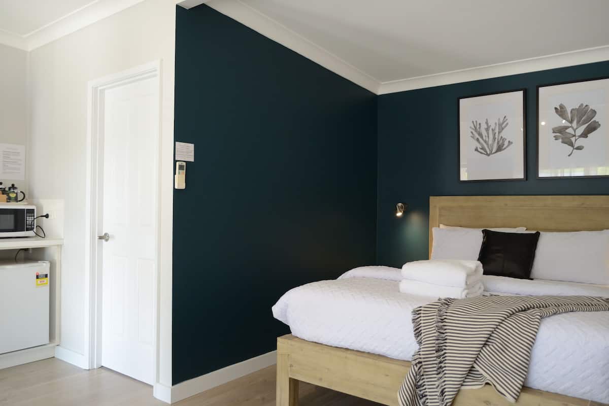A bedroom with a bed, a wooden headboard, a white bedside table with a microwave, and two framed botanical prints on the wall.