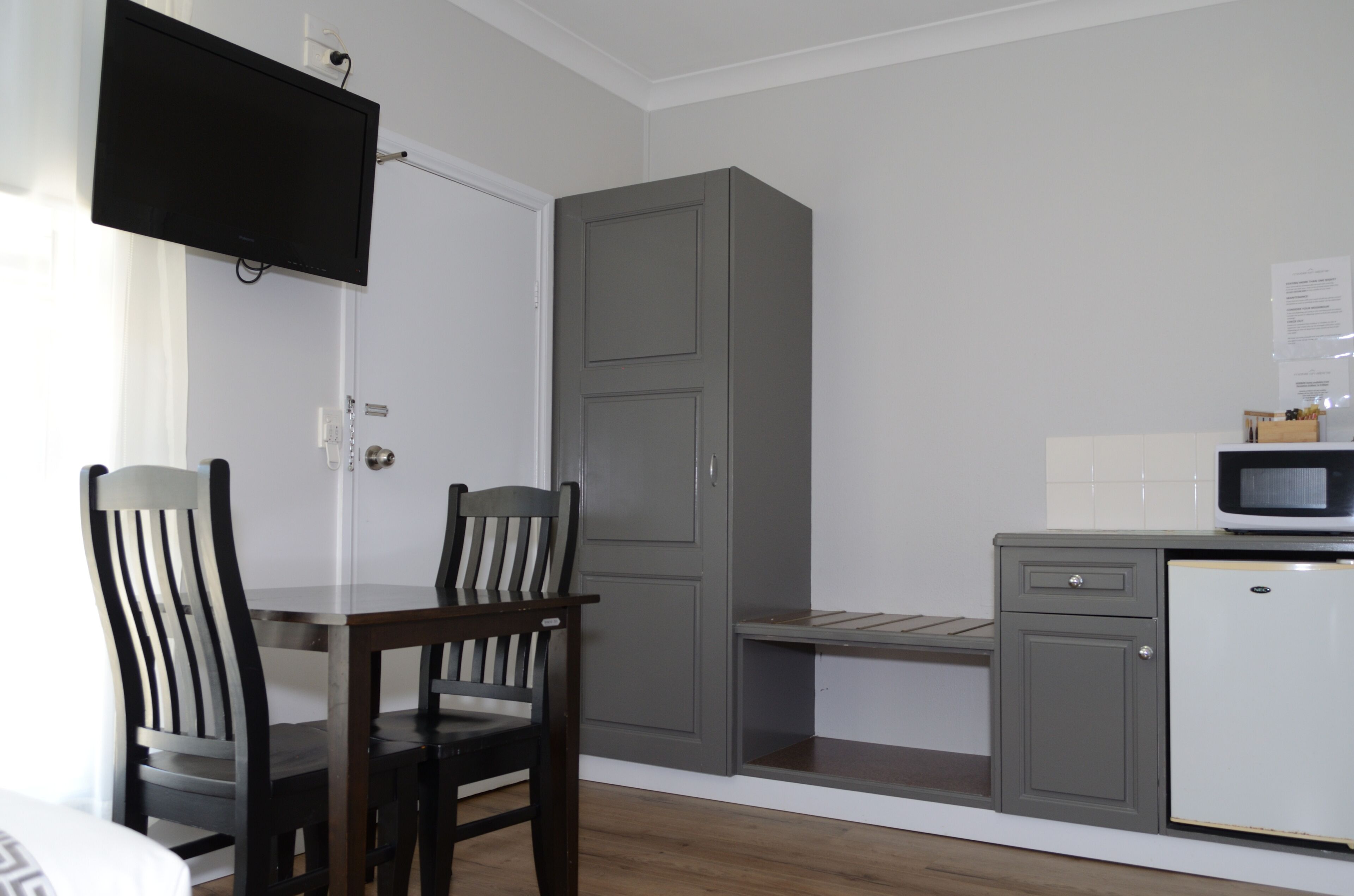 A compact room with a dining table and chairs, a grey wardrobe, a small kitchenette with a microwave, and a TV mounted on the wall.