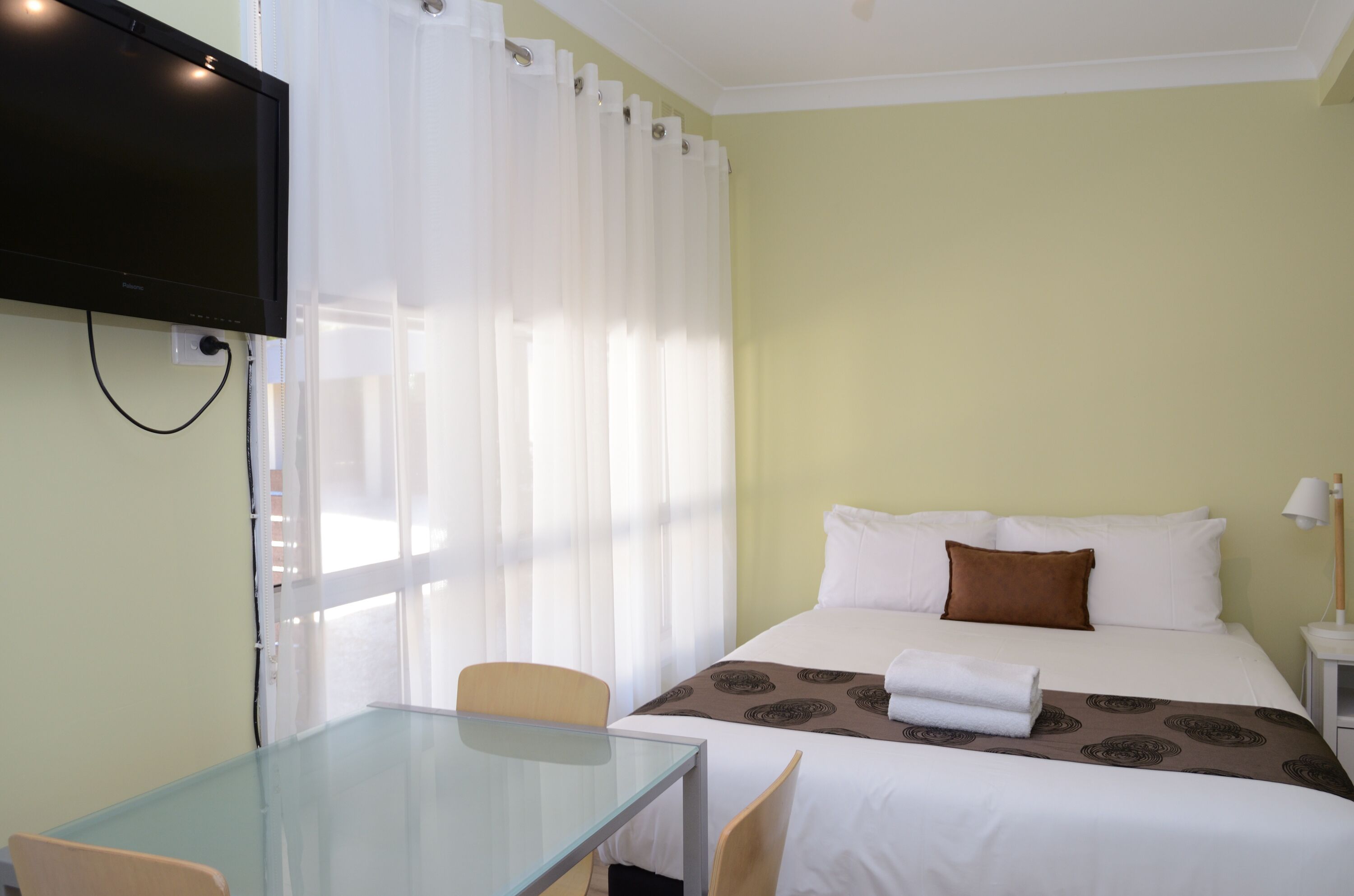 Una habitación de hotel con una cama, un televisor montado en la pared, una mesita de vidrio y una silla.