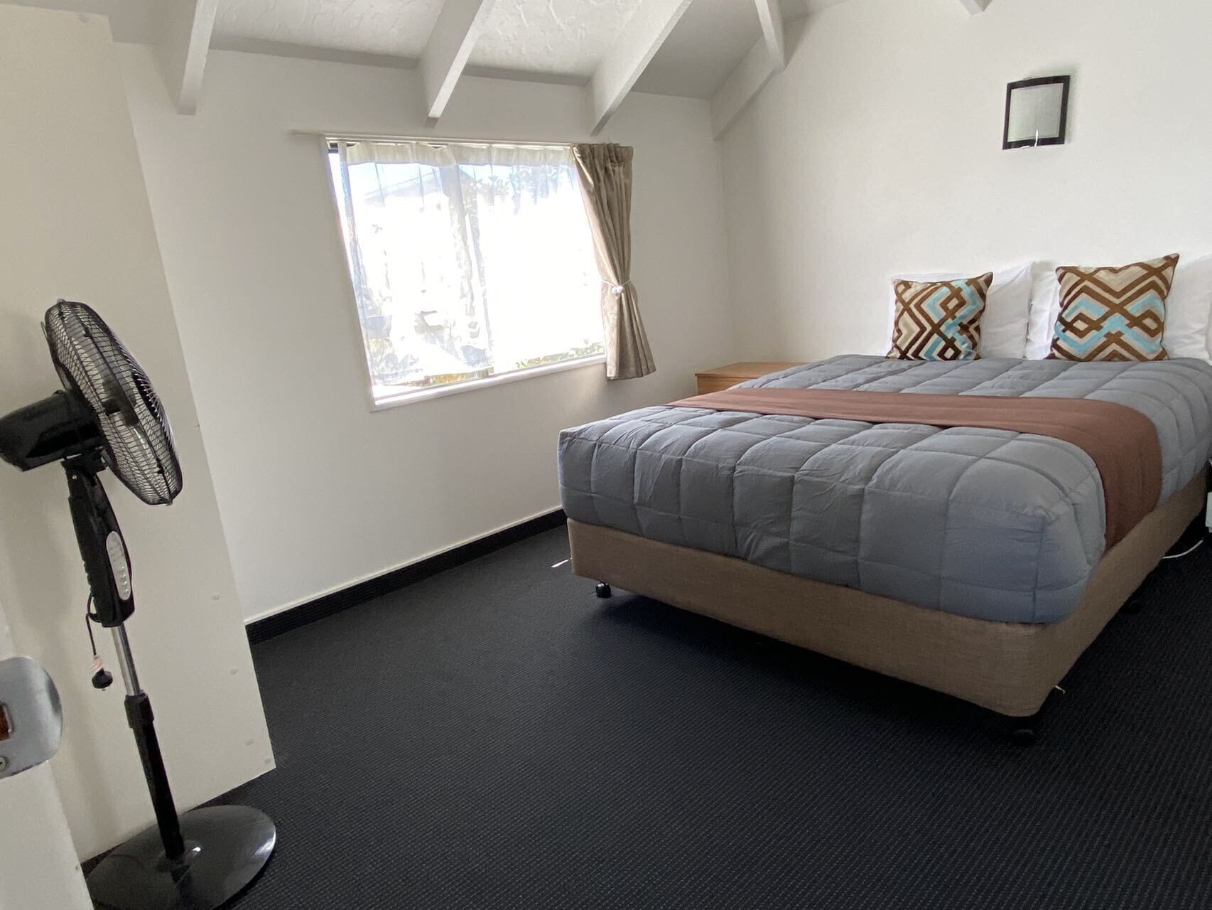 A bedroom with a bed, a standing fan, a window with curtains, and a small bedside table.