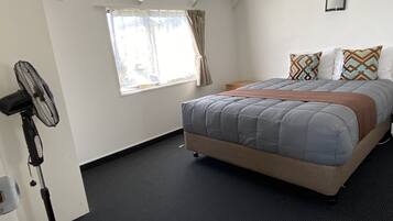 A bedroom with a bed, a standing fan, a window with curtains, and a small bedside table.