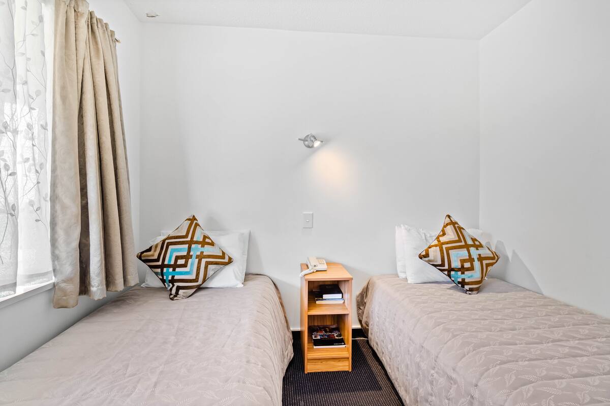 Hotellihuone, jossa on kaksi sänkyä, pieni puinen yöpöytä, jossa on puhelin ja kirjoja, sekä seinään kiinnitetty valaisin.