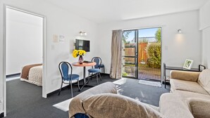 A modern hotel room with a bed, a sofa, a small table with chairs, a TV, and a sliding glass door to the outside. - Ascot Vale Motor Lodge (Christchurch)