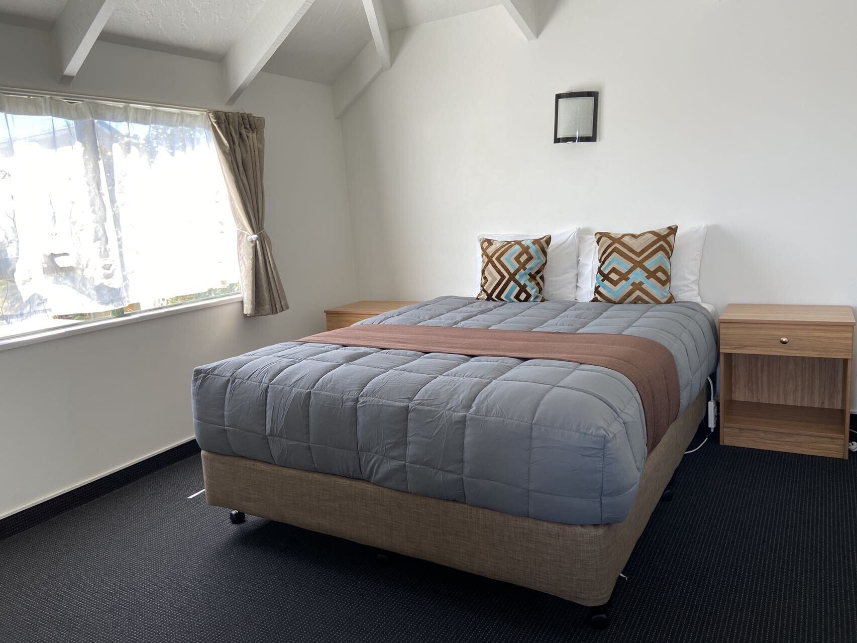 Quarto bem arrumado com cama, mesas de cabeceira, janela com cortinas e luminária na parede.