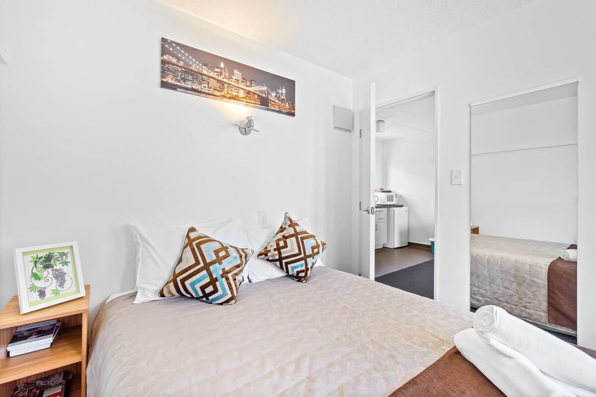 Hotellihuone, jossa on sänky, yöpöytä, jossa on kehystetty kuva, seinällä oleva kaupunkimaisemaa esittävä taideteos ja avoimesta ovesta näkyvä kylpyhuone.