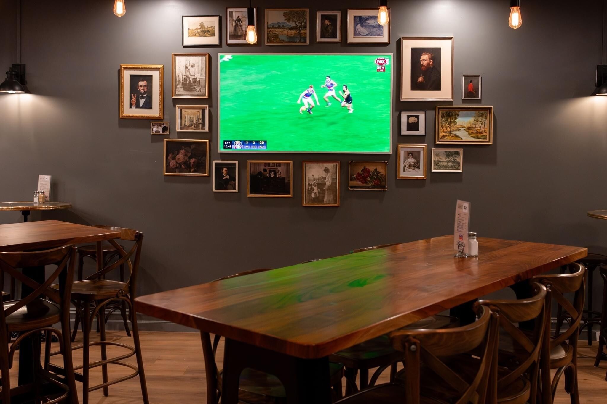 Quarto com uma televisão grande exibindo um jogo esportivo, obras de arte nas paredes e mobiliário de madeira.