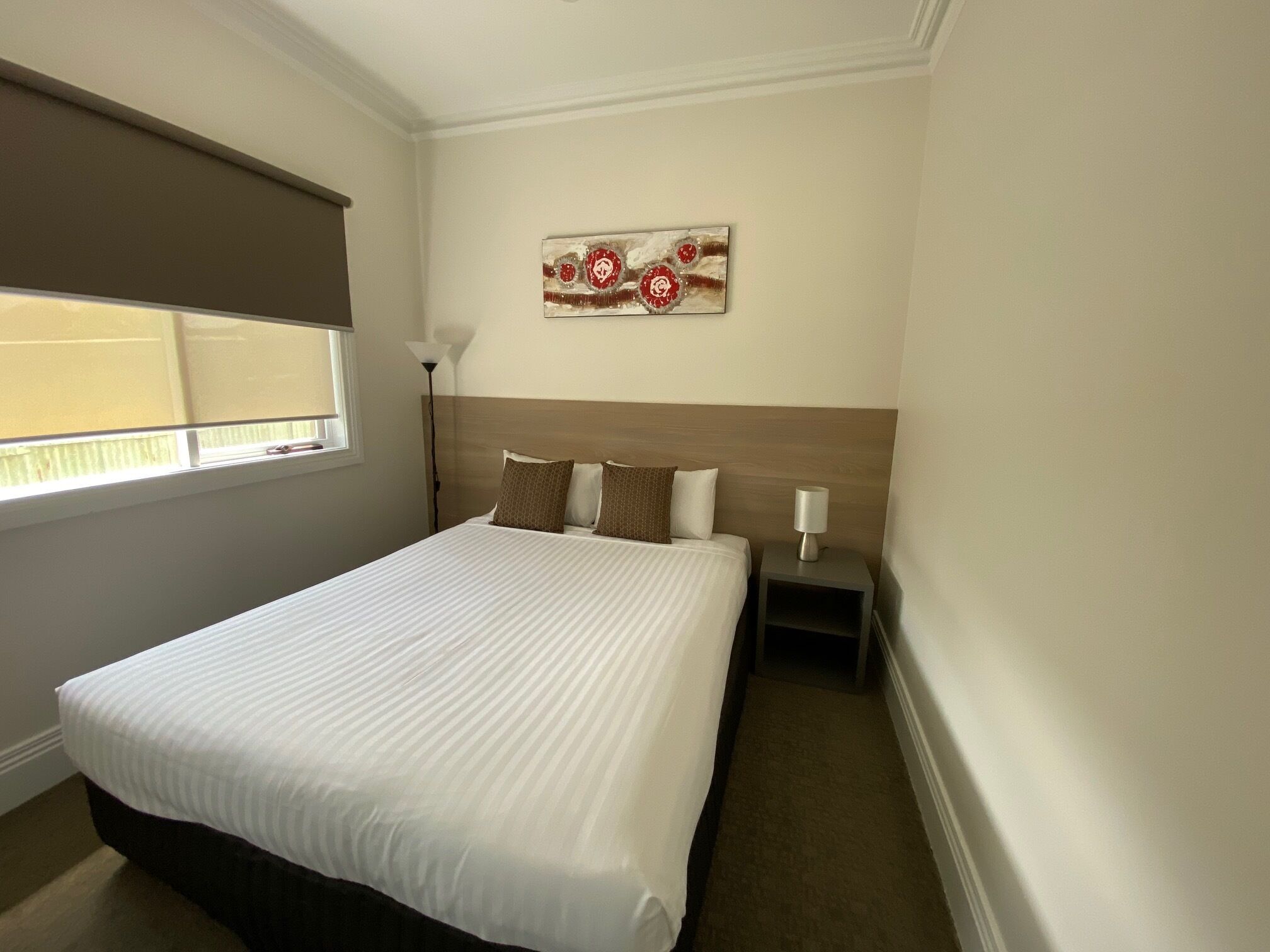 A hotel room with a large bed, a nightstand with a lamp, a window with blinds, and a wall art piece.