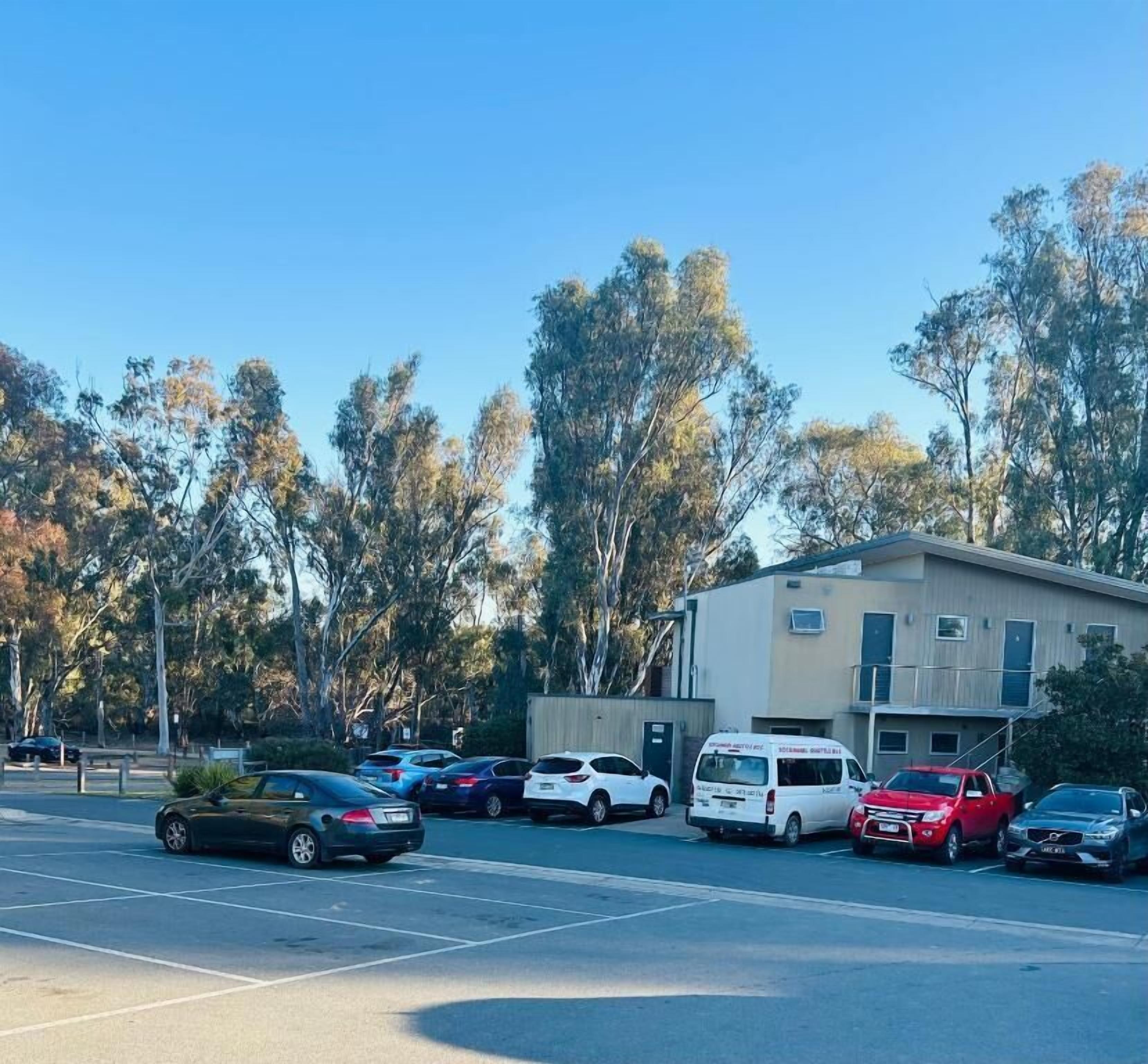 Un estacionamiento con varios coches y un edificio de techo plano y múltiples ventanas.