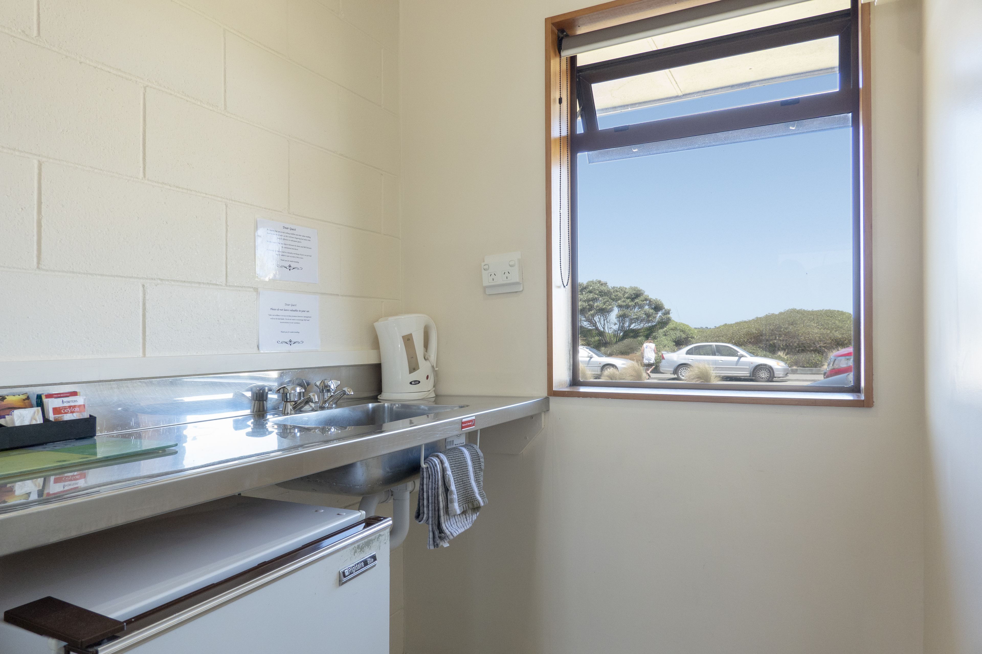 Una pequeña kitchenette con un lavamanos, una tetera y una ventana con vistas a un estacionamiento.