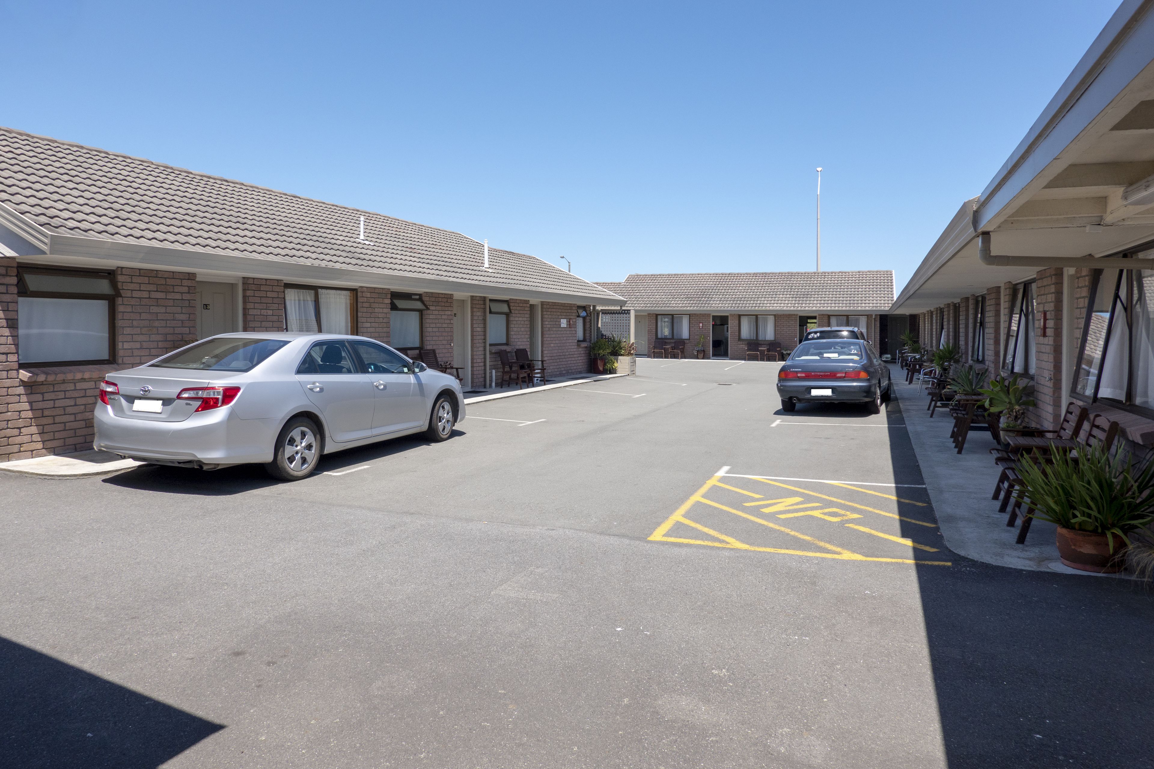 A motel with two-story brick buildings, a parking lot, and a row of potted plants along the sidewalk.