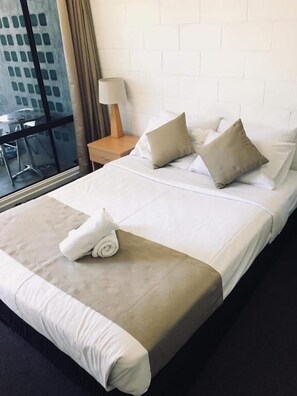 A hotel room with a large bed, two pillows, a bedside table with a lamp, and a view of the outside through a window.