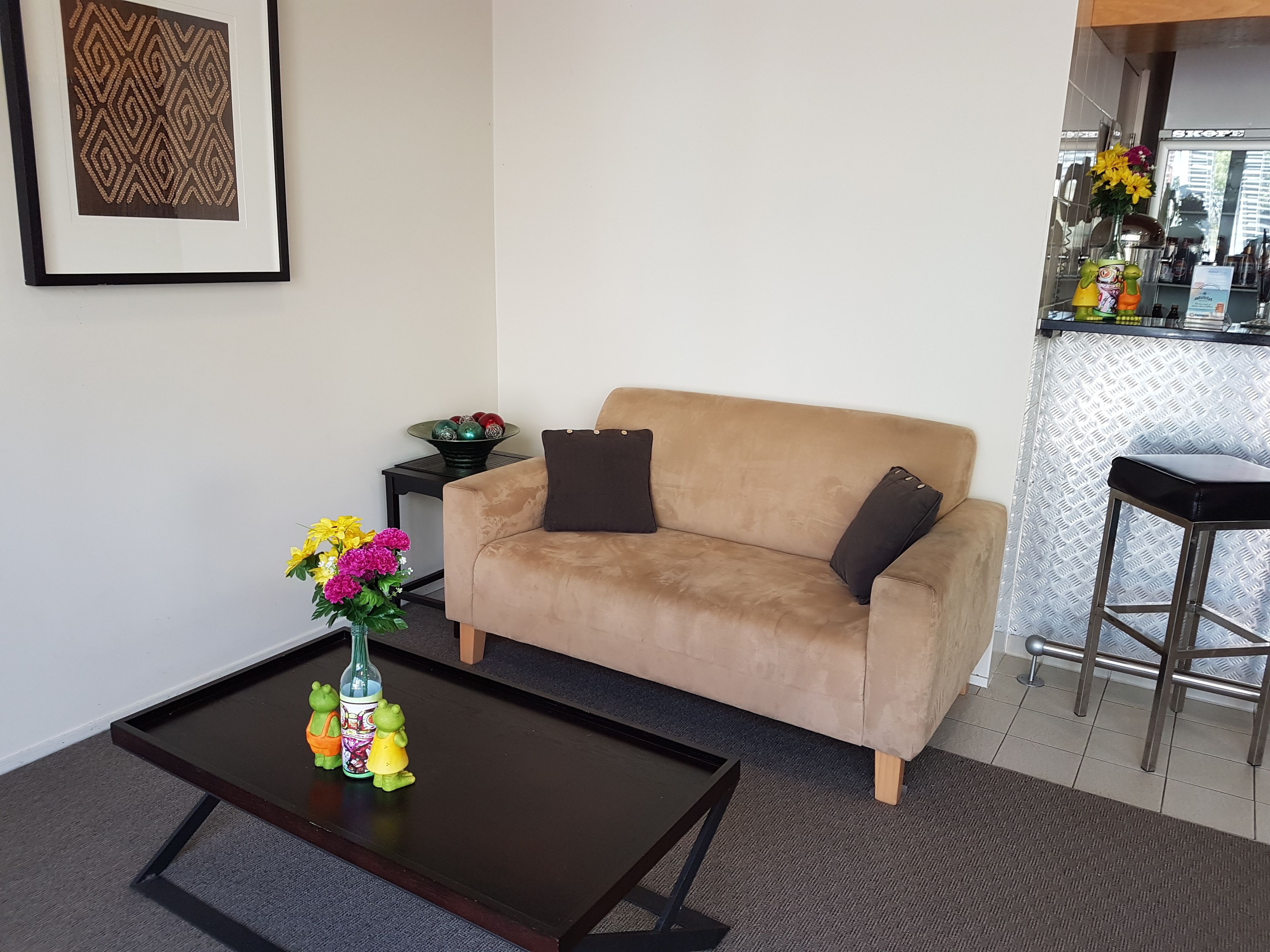 A living room with a beige sofa, a small side table, a coffee table, and a kitchen area with a bar stool.