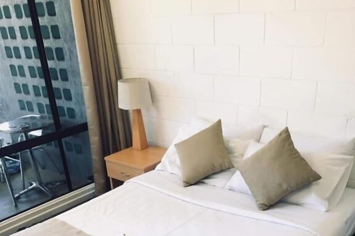 A hotel room with a bed, a nightstand, a lamp, and a window with a view of a building.