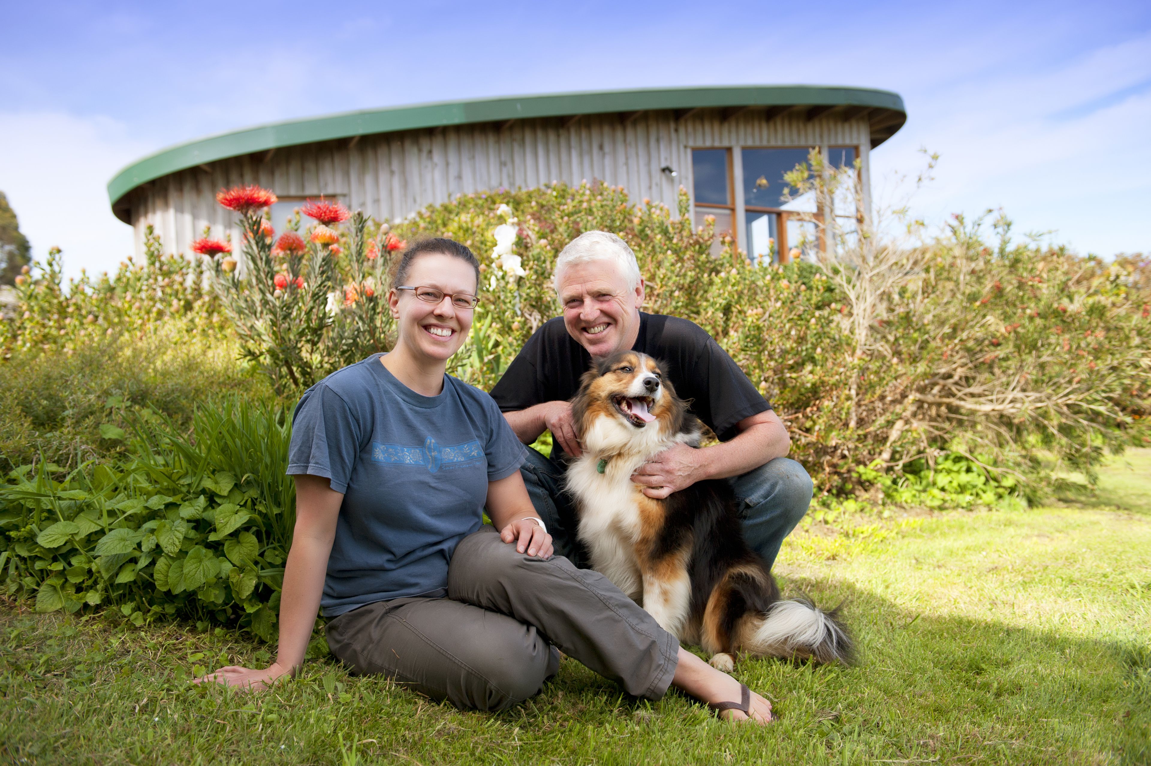 Two people sitting on grass with a dog in front of a building with a curved roof and windows.