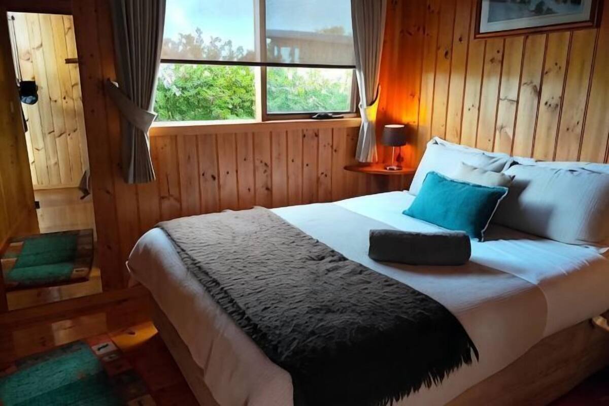 Quarto com painéis de madeira, uma cama, uma janela e uma mesa de cabeceira com abajur.