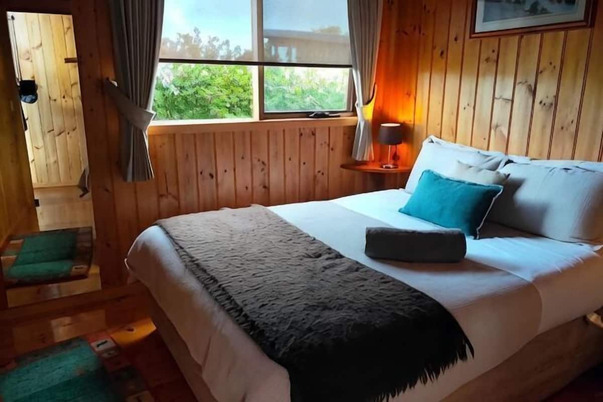 A wooden-paneled room with a bed, a window, and a nightstand with a lamp.