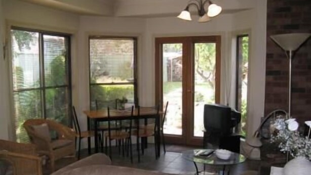 A living room with a sofa, a dining table with chairs, a television, and a lamp. There are large windows and a door leading outside. - Beechworth Gorge Walk Guest House (Beechworth)