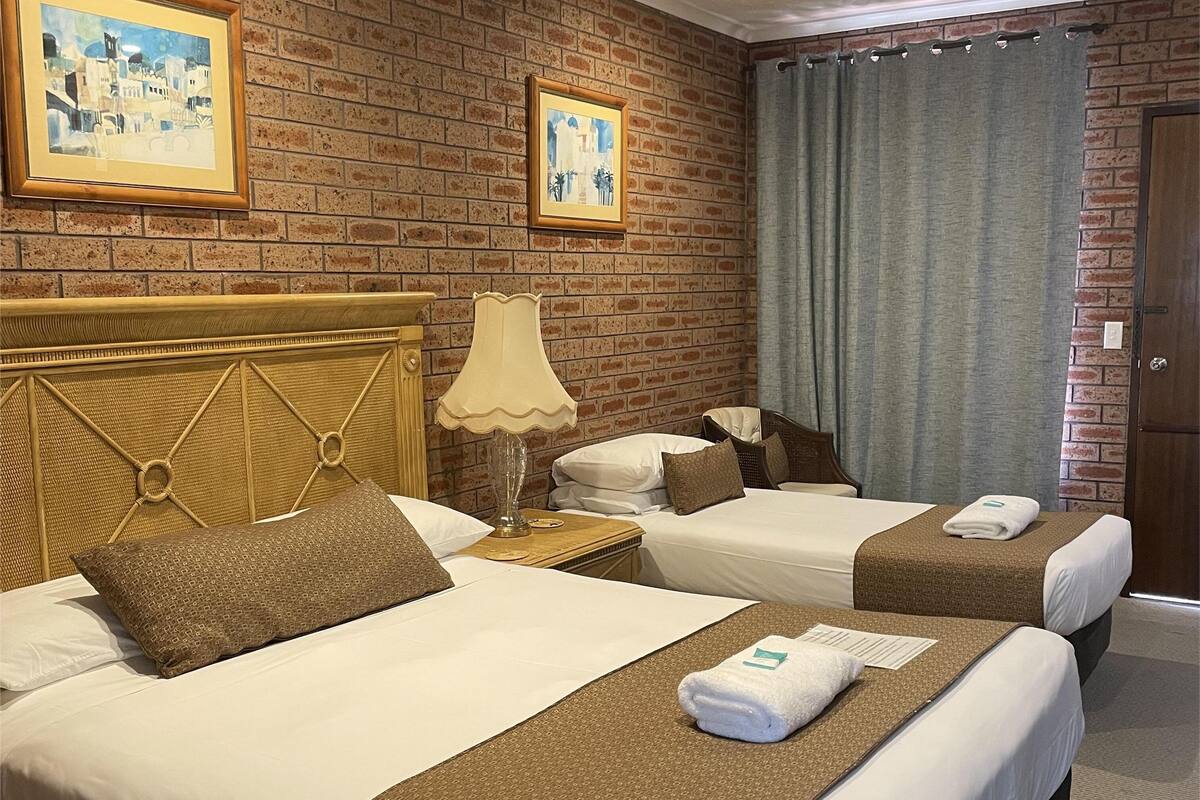 Ein Hotelzimmer mit zwei Betten, einem hölzernen Kopfteil, einer Lampe und zwei gerahmten Bildern an der Ziegelwand.