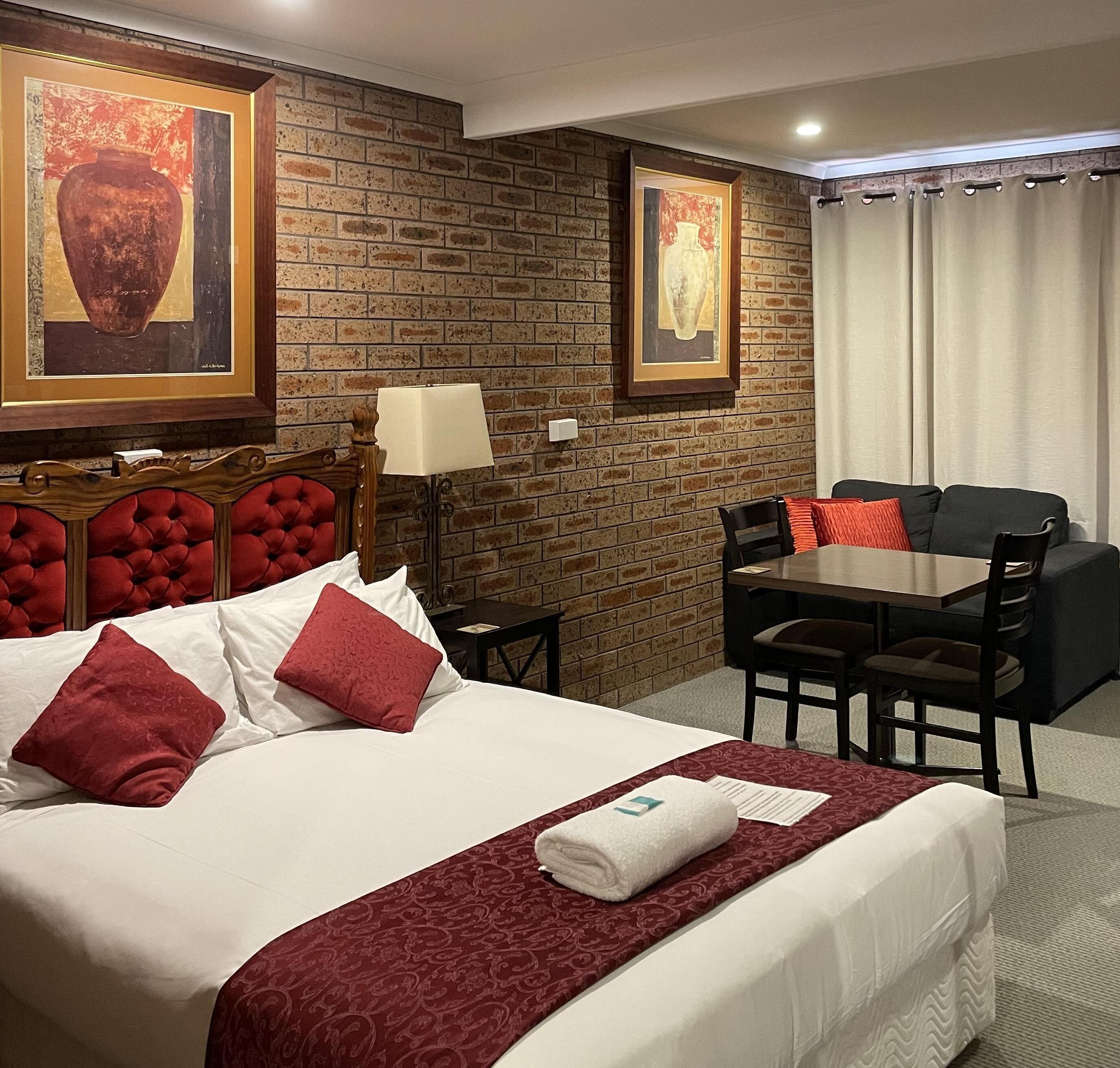 Habitación de hotel con pared de ladrillo, una cama grande con ropa de cama roja y blanca, cabecera, mesa con sillas y una lámpara.