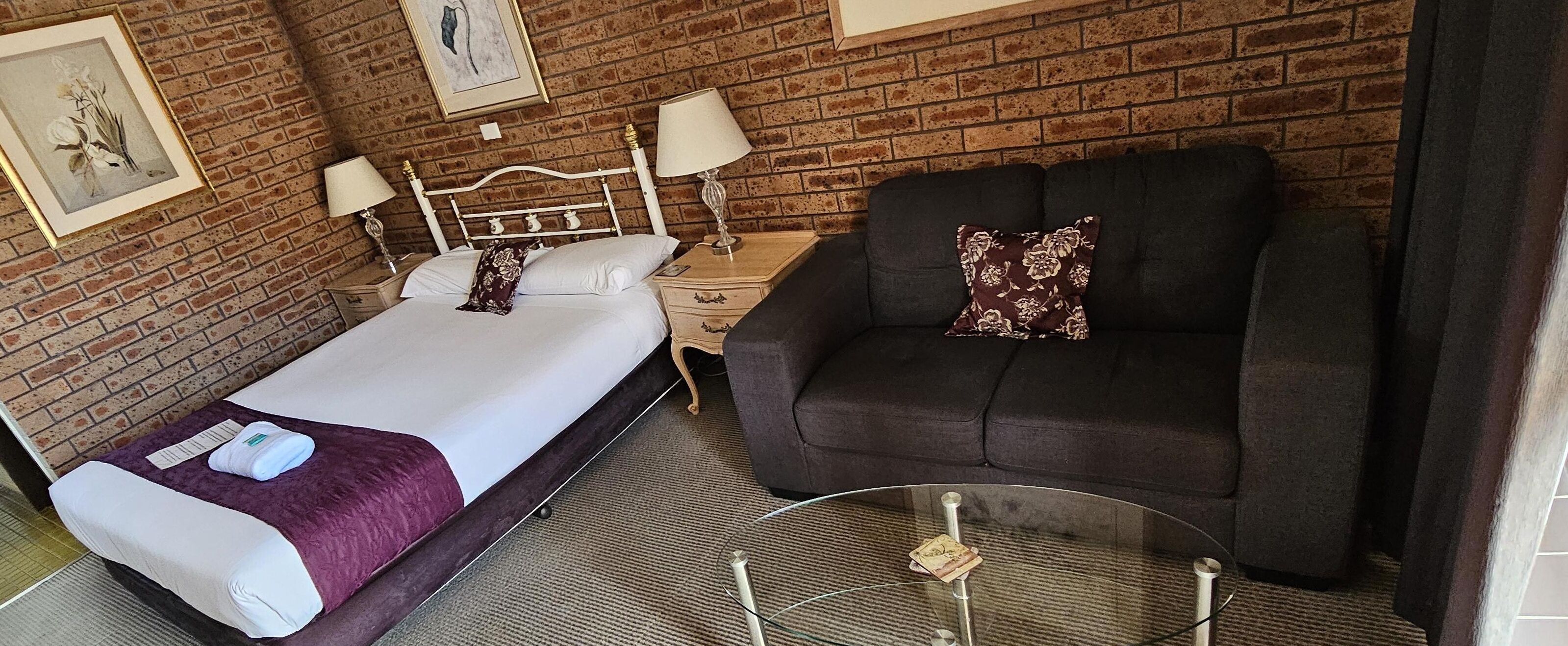 A hotel room with a bed, a sofa, a glass coffee table, and a brick wall.