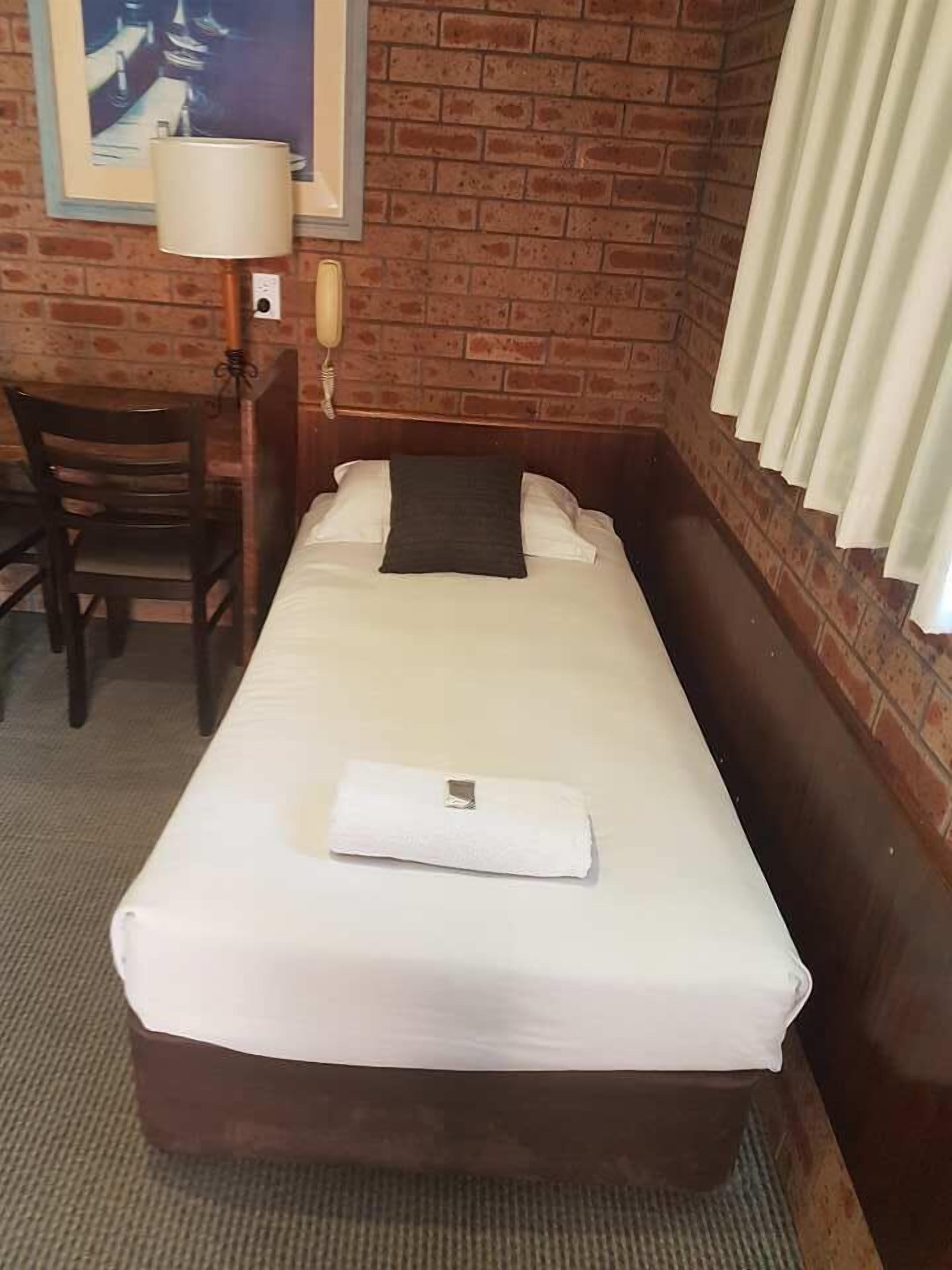 A single bed with a white mattress and pillow, a wooden headboard, a small table with a chair, a lamp, and a framed picture on the wall.