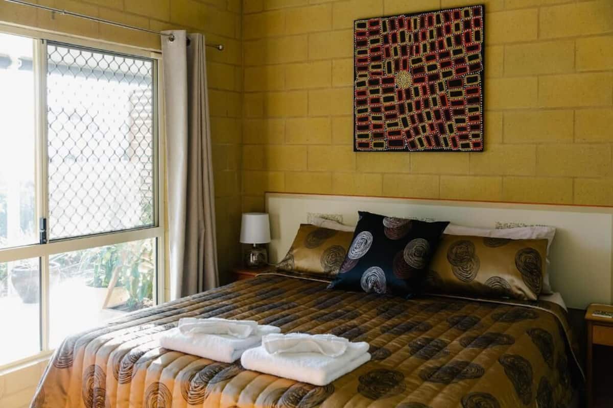 A hotel room with a large bed, a window with a view, a wall art piece, and a nightstand with a lamp.