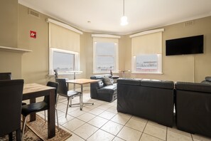A living room with a sofa, a dining table with chairs, a TV mounted on the wall, and large windows.