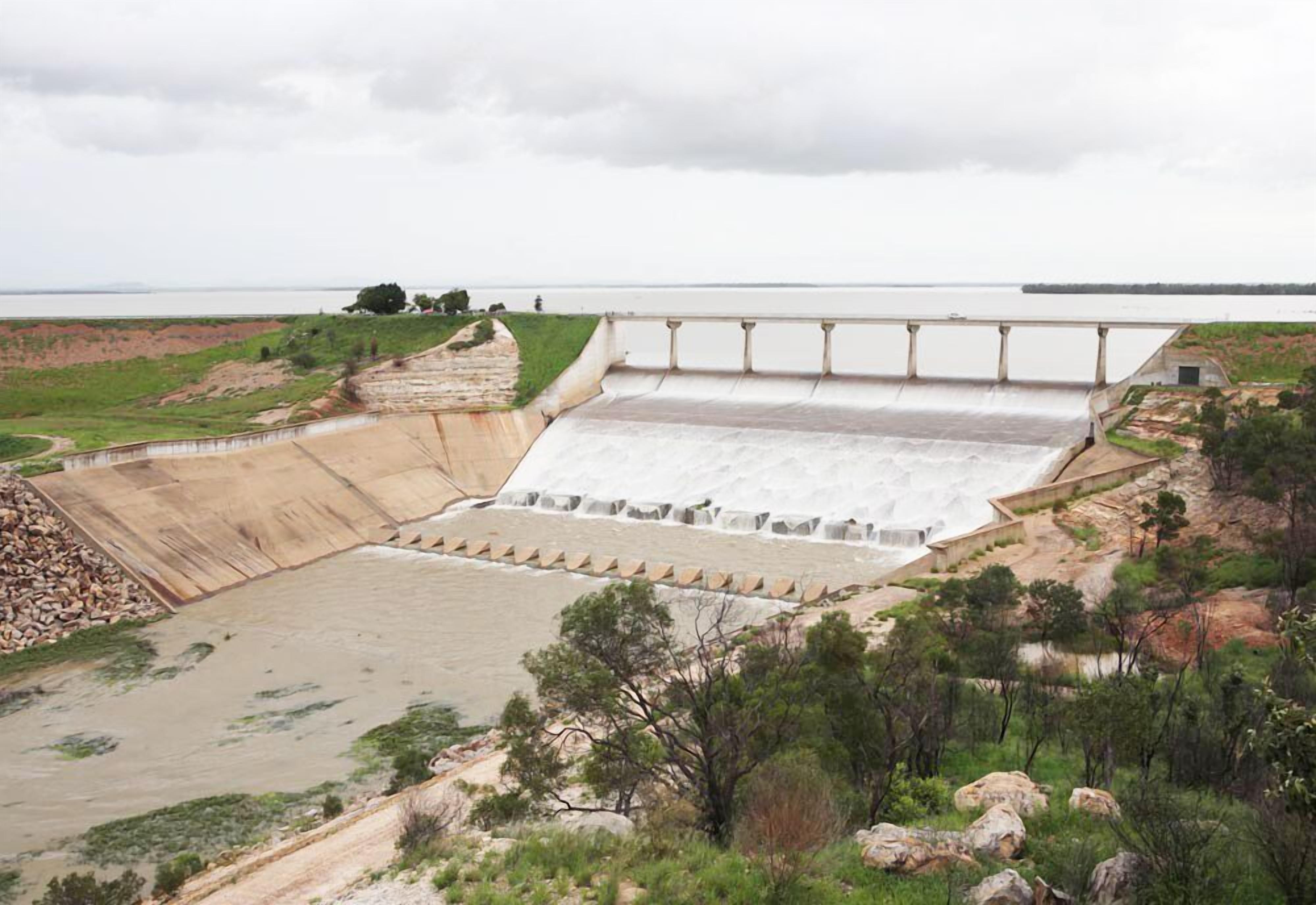 A large dam with a spillway, surrounded by rocky terrain and sparse vegetation.