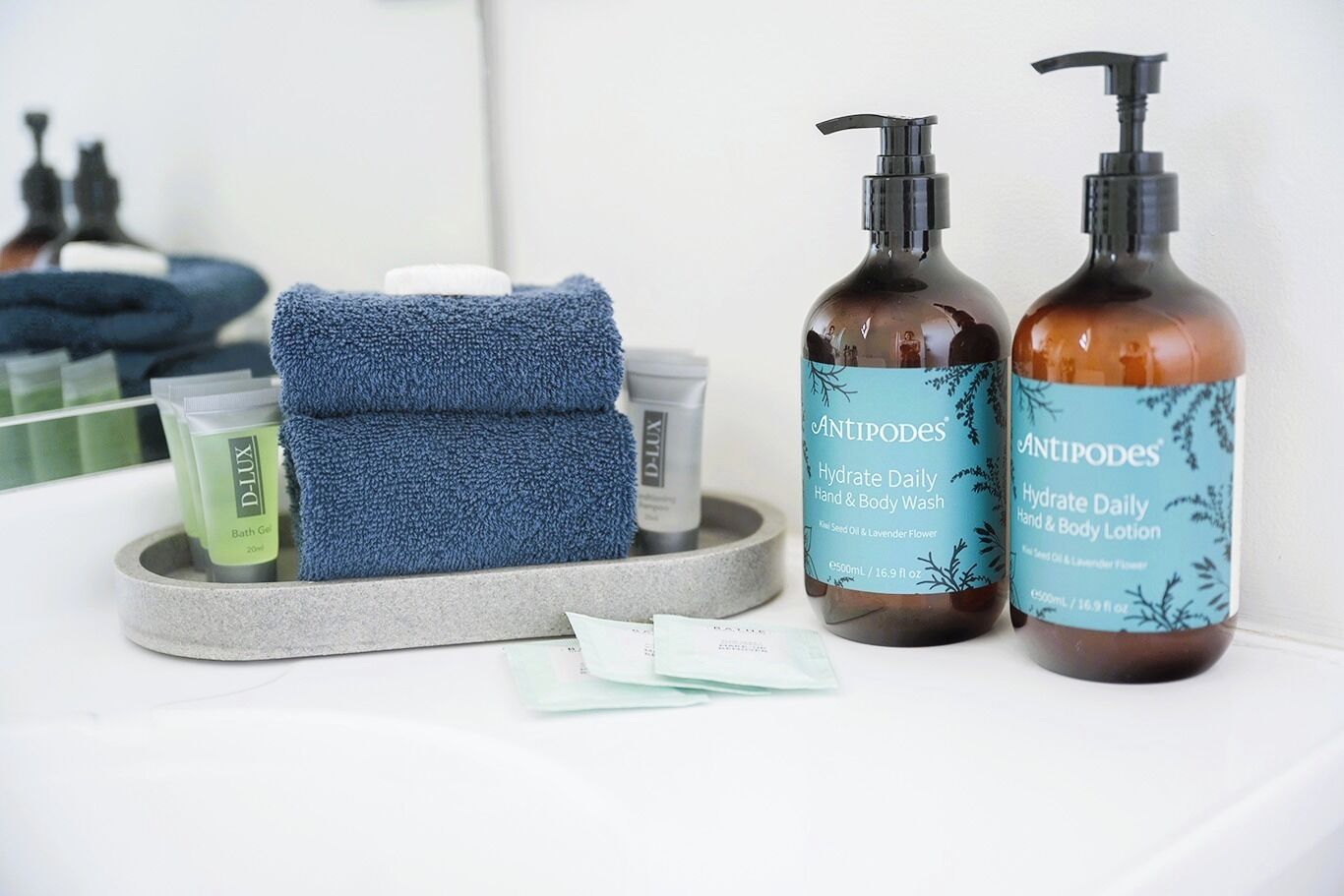 Un baño con una toalla azul, dos botellas de lavamanos y gel corporal Antipodes, un gel de baño verde y una pequeña caja de pañuelos.