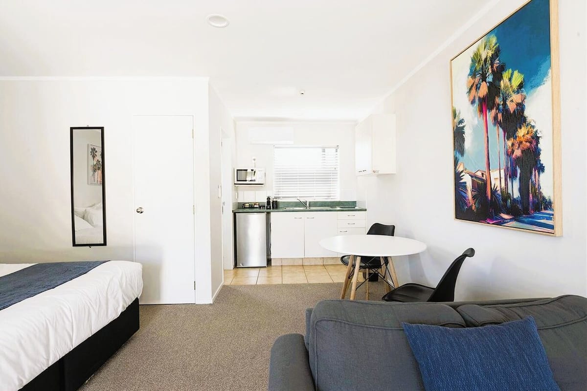A hotel room with a bed, a sofa, a dining table with chairs, a kitchenette, and a painting on the wall.